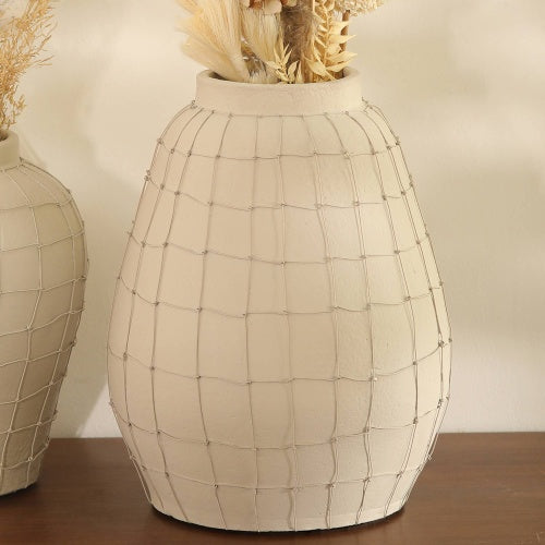 A white ceramic vase with a grid pattern, filled with dried beige pampas grass and other dried plants, placed on a wooden surface against a light-colored wall.