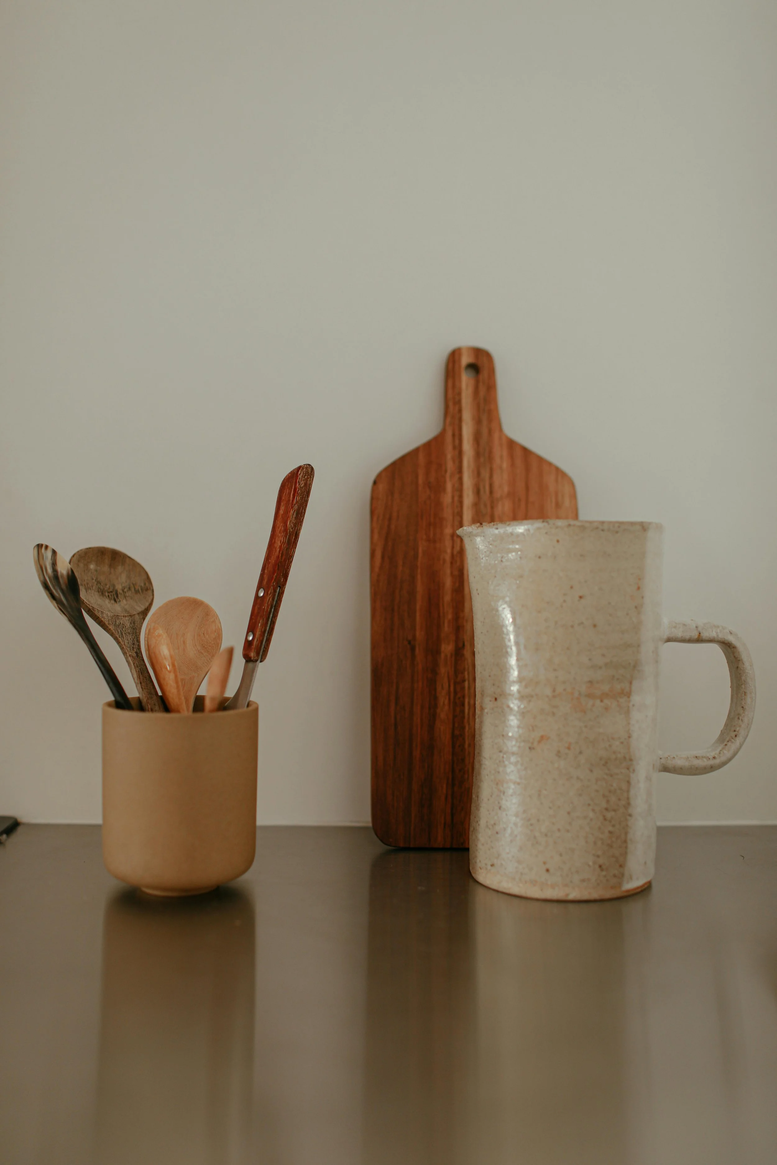 Kitchen countertop with a beige cup holding wooden and metal spoons, a wooden cutting board, and a speckled ceramic mug.