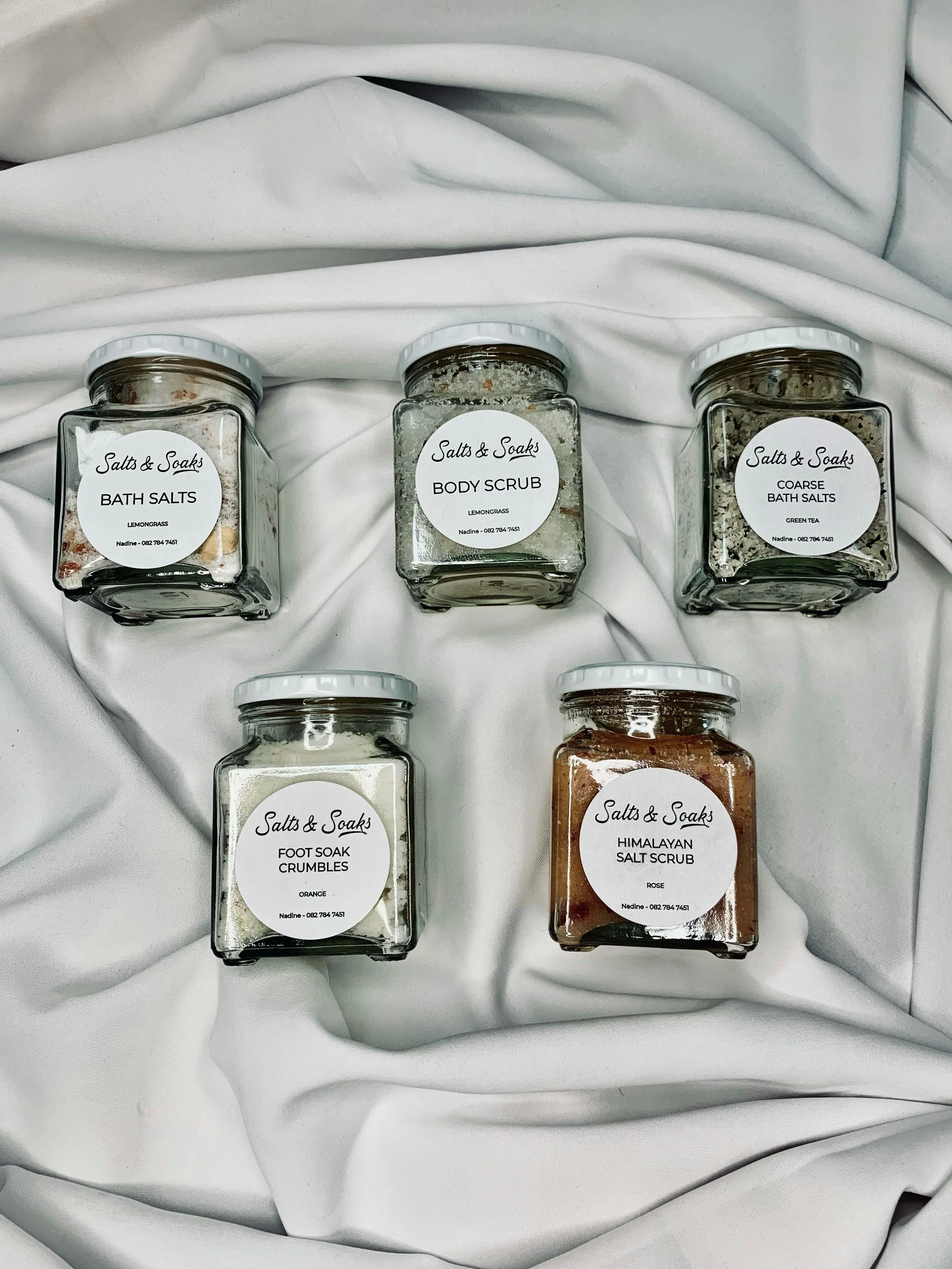 Five jars of bath and body salts on a white fabric background, labeled with different scents including lemongrass, green tea, orange, rose, and a body scrub in the center.