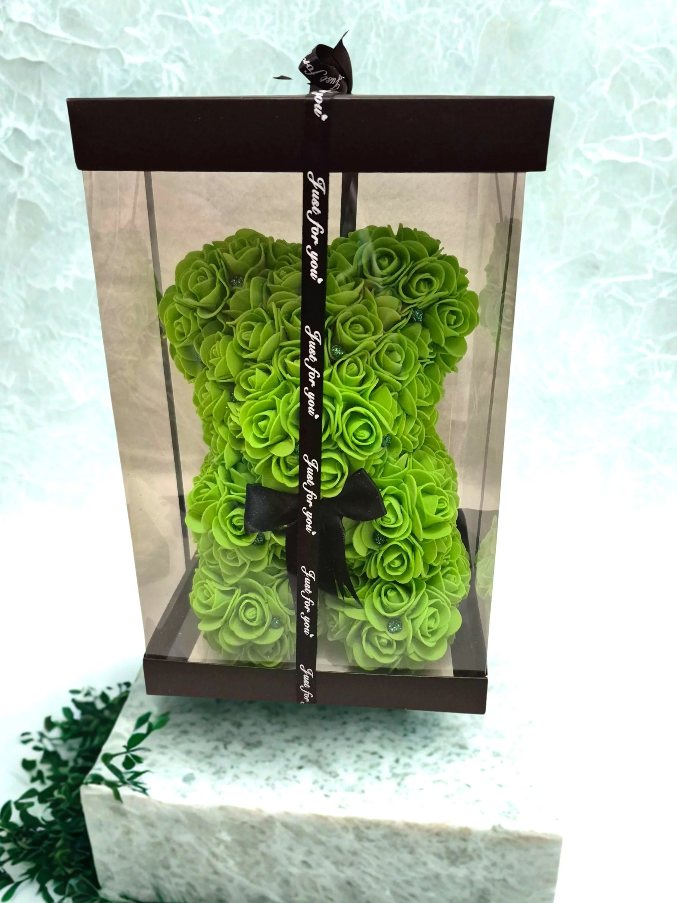 Green teddy bear made of artificial roses in a clear gift box with a black ribbon.