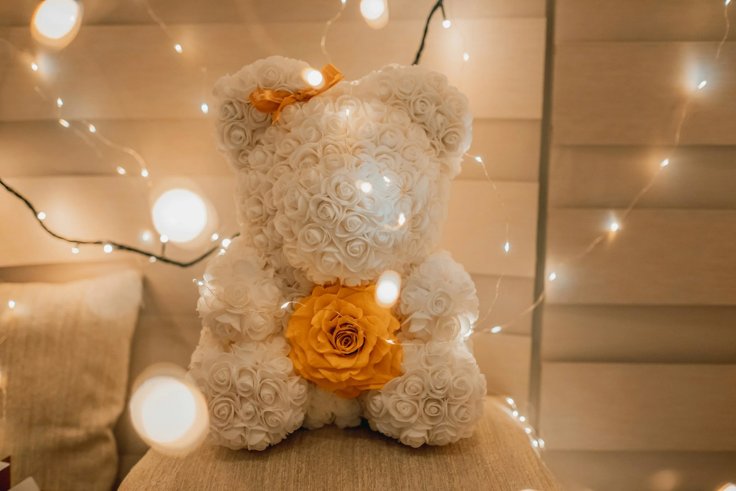 A teddy bear made of white roses, with a yellow rose in its lap and a yellow rose on its head, surrounded by string lights, against a wooden wall background.
