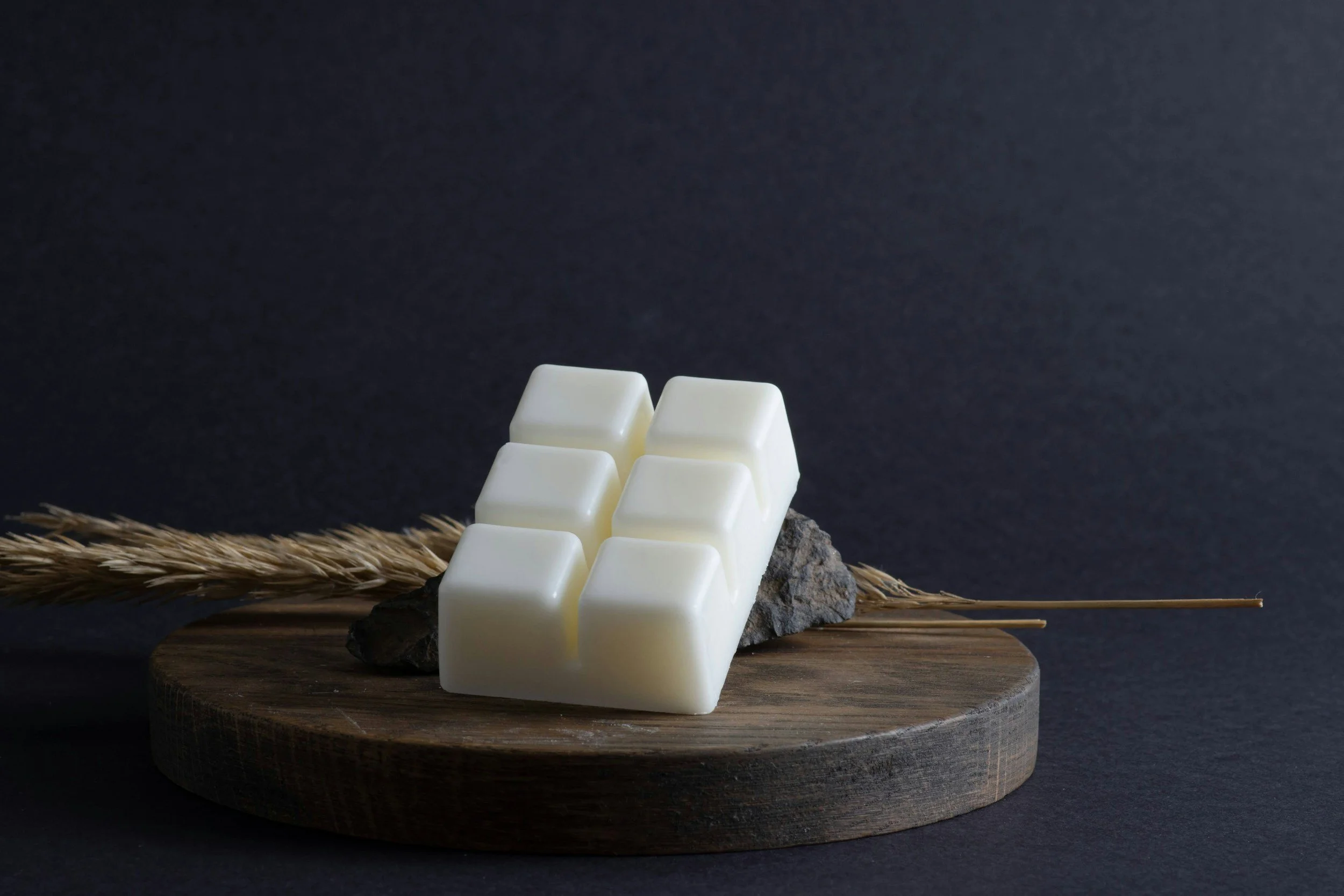 White wax cubes on a dark wooden board with a wheat stalk, set against a dark background.