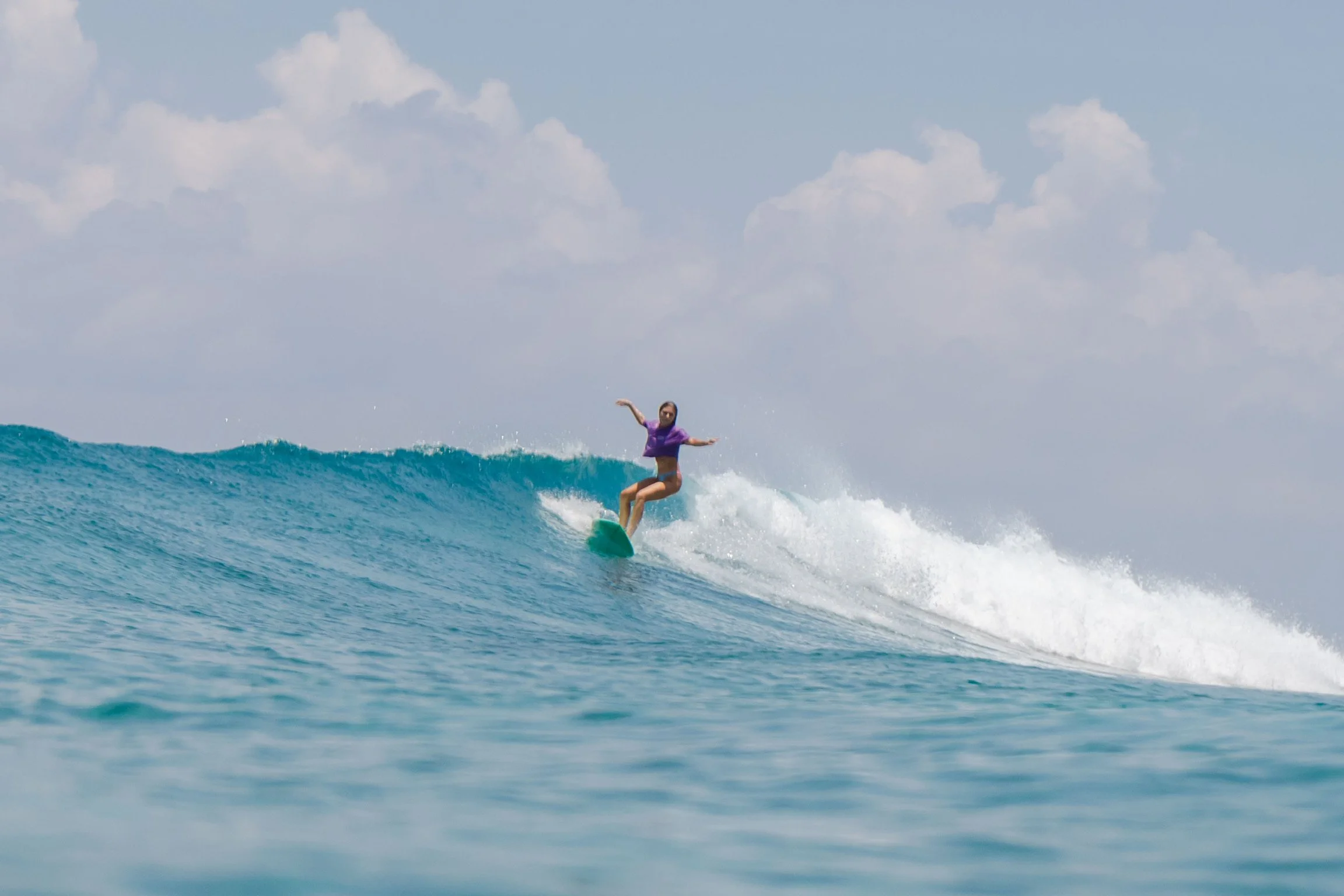 A woman surfing on a wave in the ocean, wearing a purple rash guard and shorts.