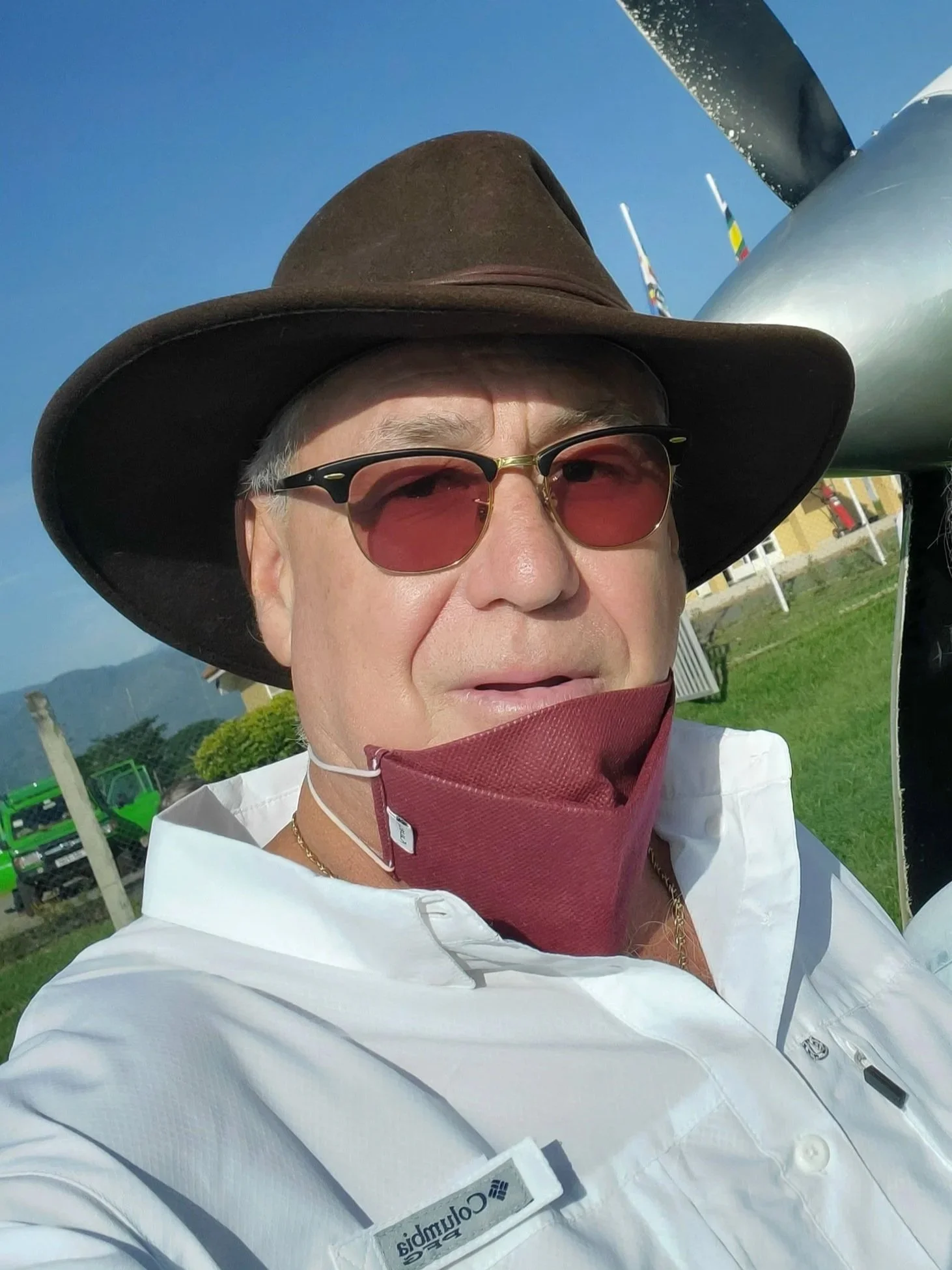 A person wearing a wide-brimmed hat, sunglasses, a white Columbia shirt, and a red face mask under their chin, standing outdoors with a blue sky, grass, and flags in the background.