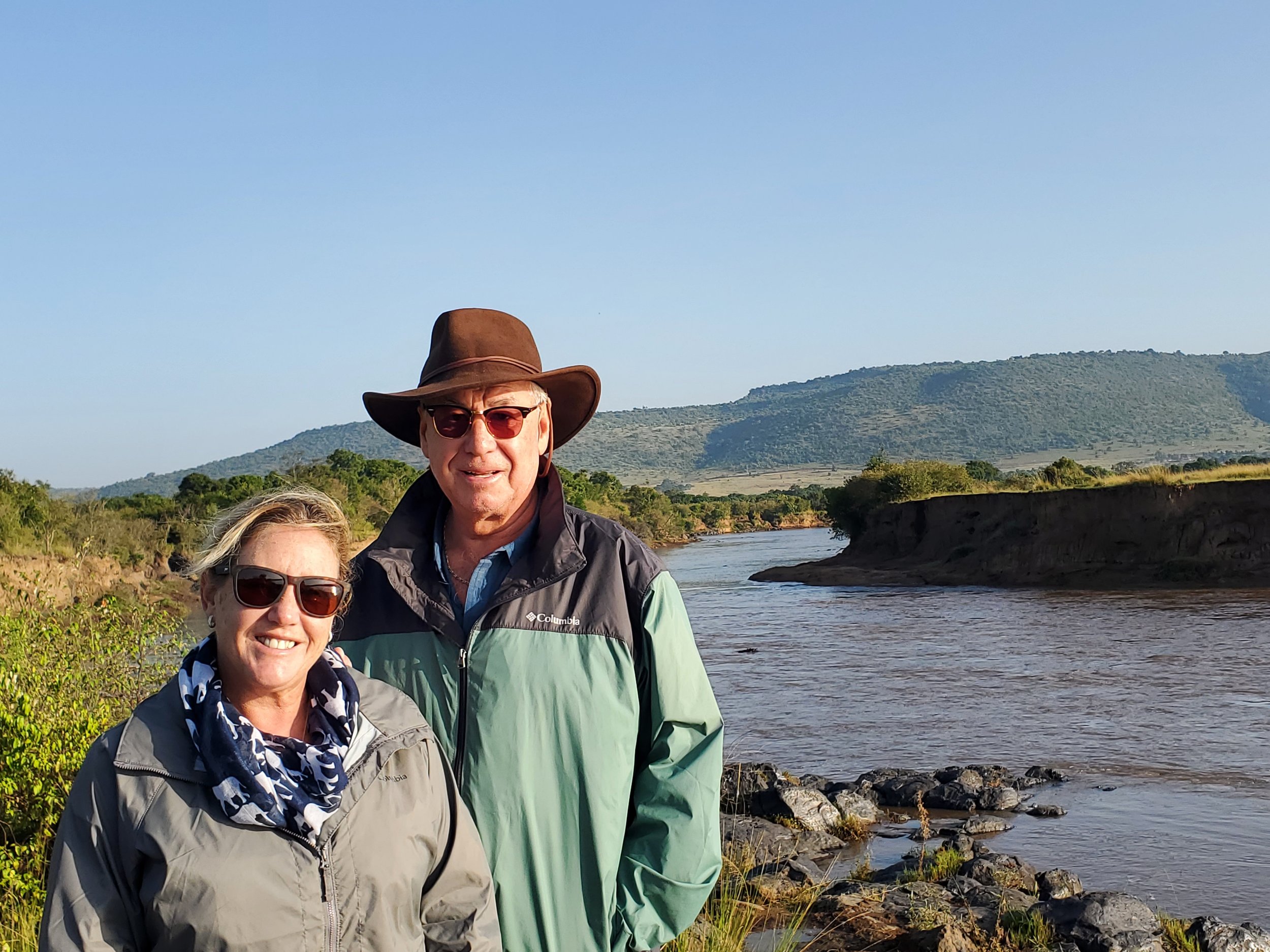 A smiling elderly couple wearing sunglasses and outdoor jackets standing beside a river with mountains in the background during daytime.