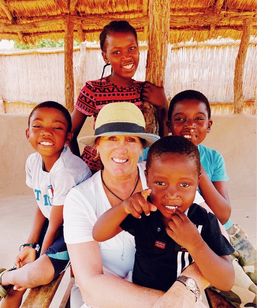 A smiling woman wearing a hat sits with four children in an outdoor setting with a thatched roof structure.