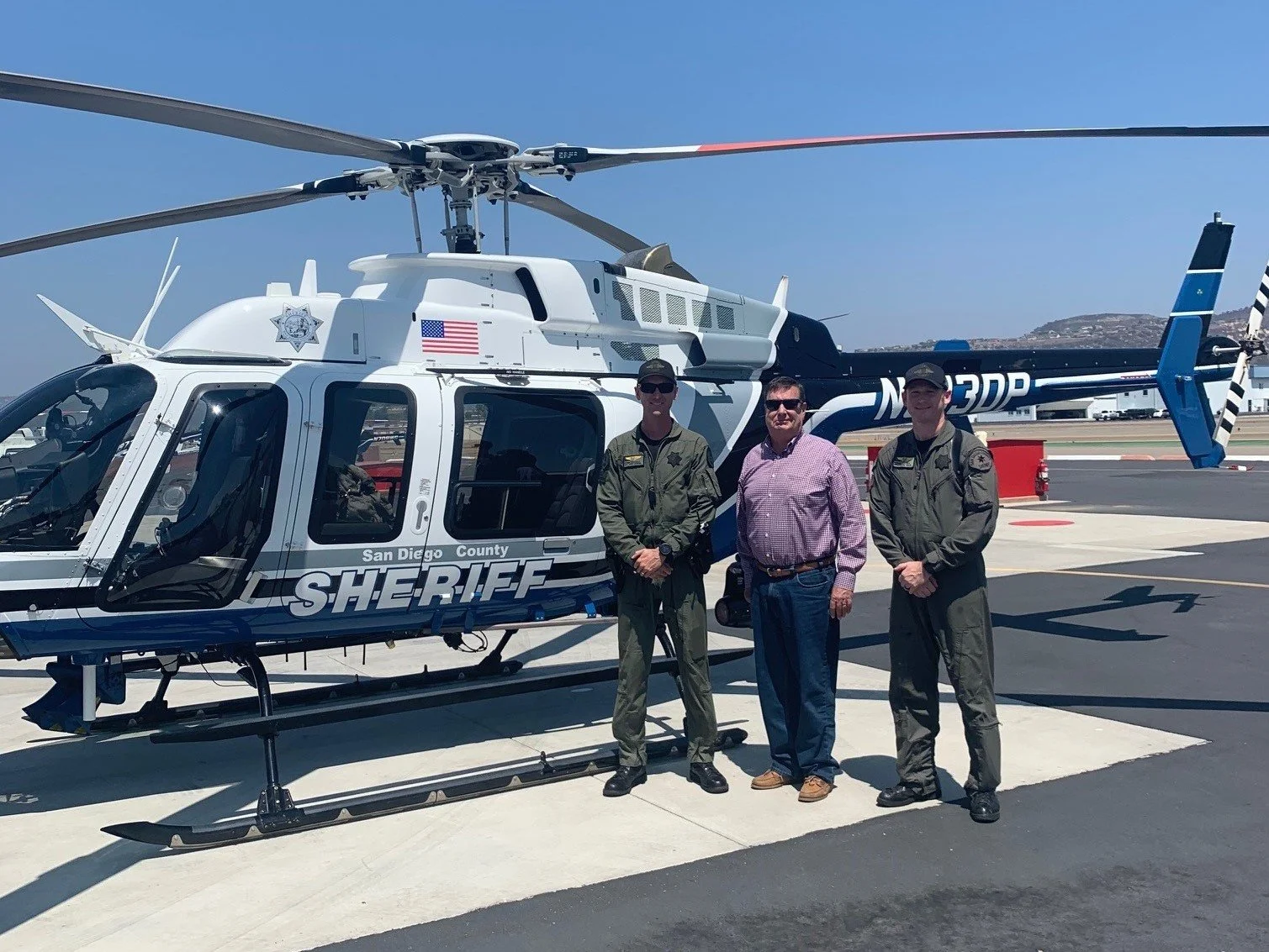 Three men standing next to a San Diego County Sheriff's helicopter on a tarmac, with clear blue skies in the background.