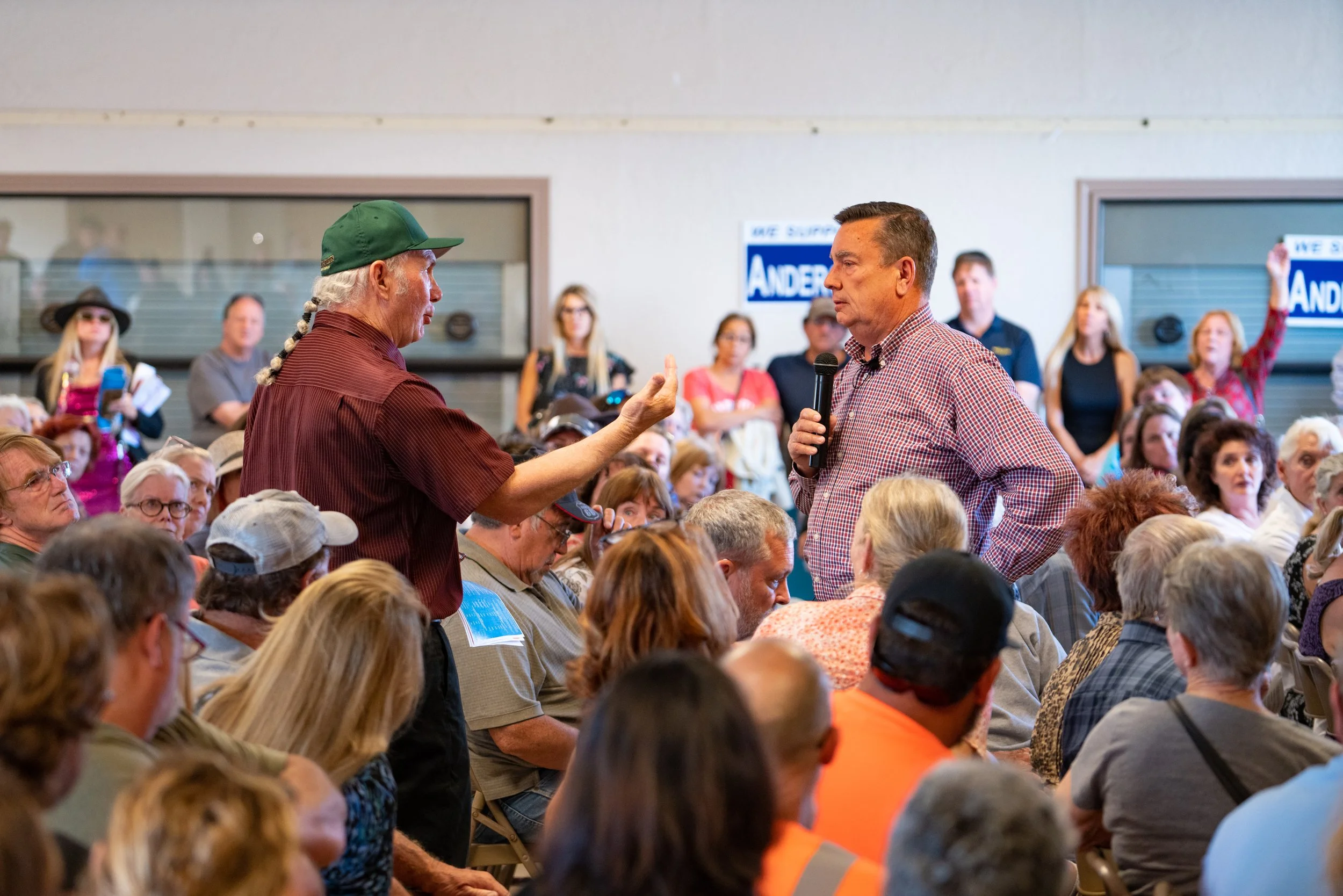 Man with gray braid and green cap speaking to man with microphone at a political rally.