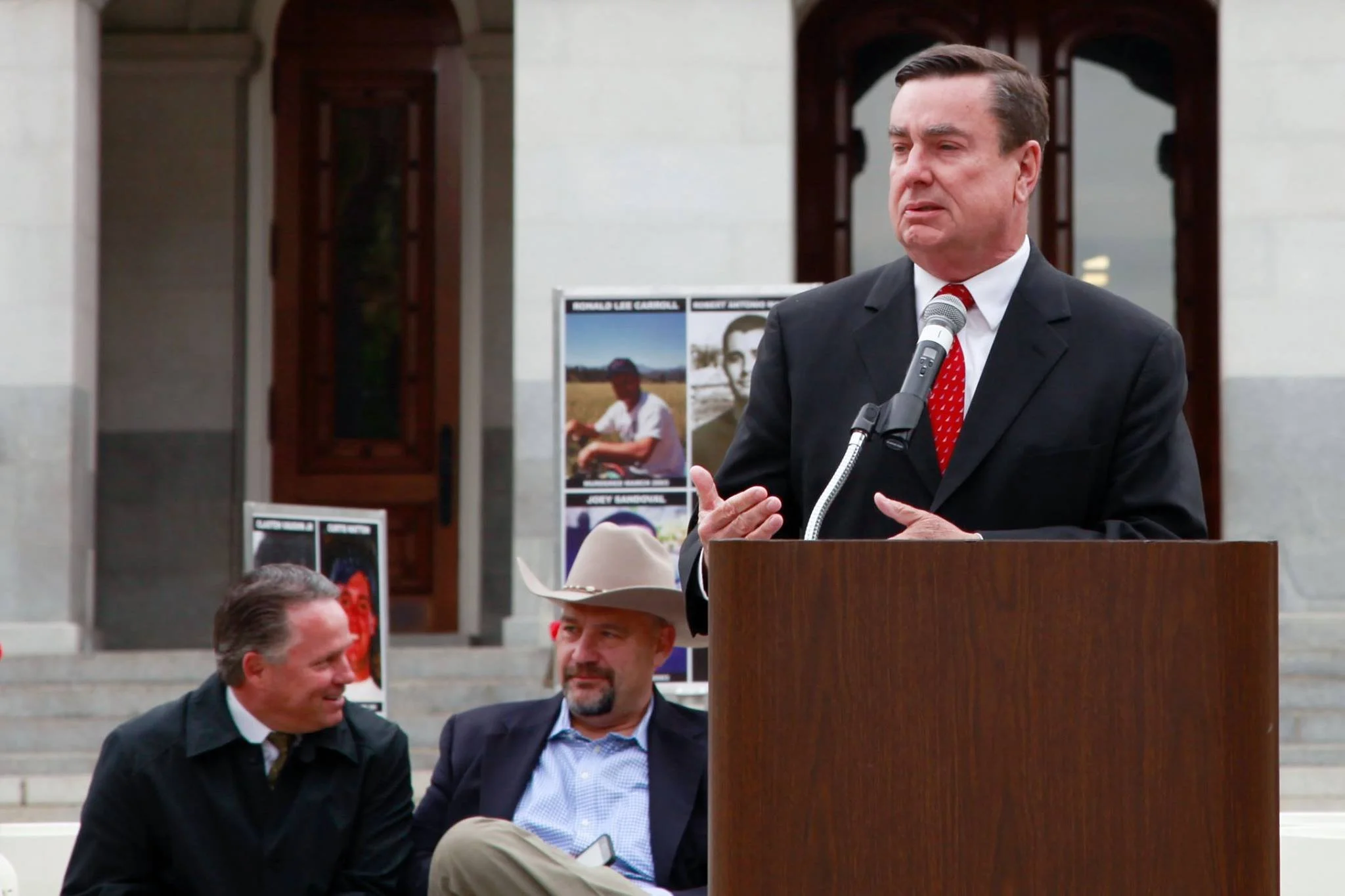 A man in a suit and red tie speaking at a podium outside a government building, with two men seated on the steps listening.