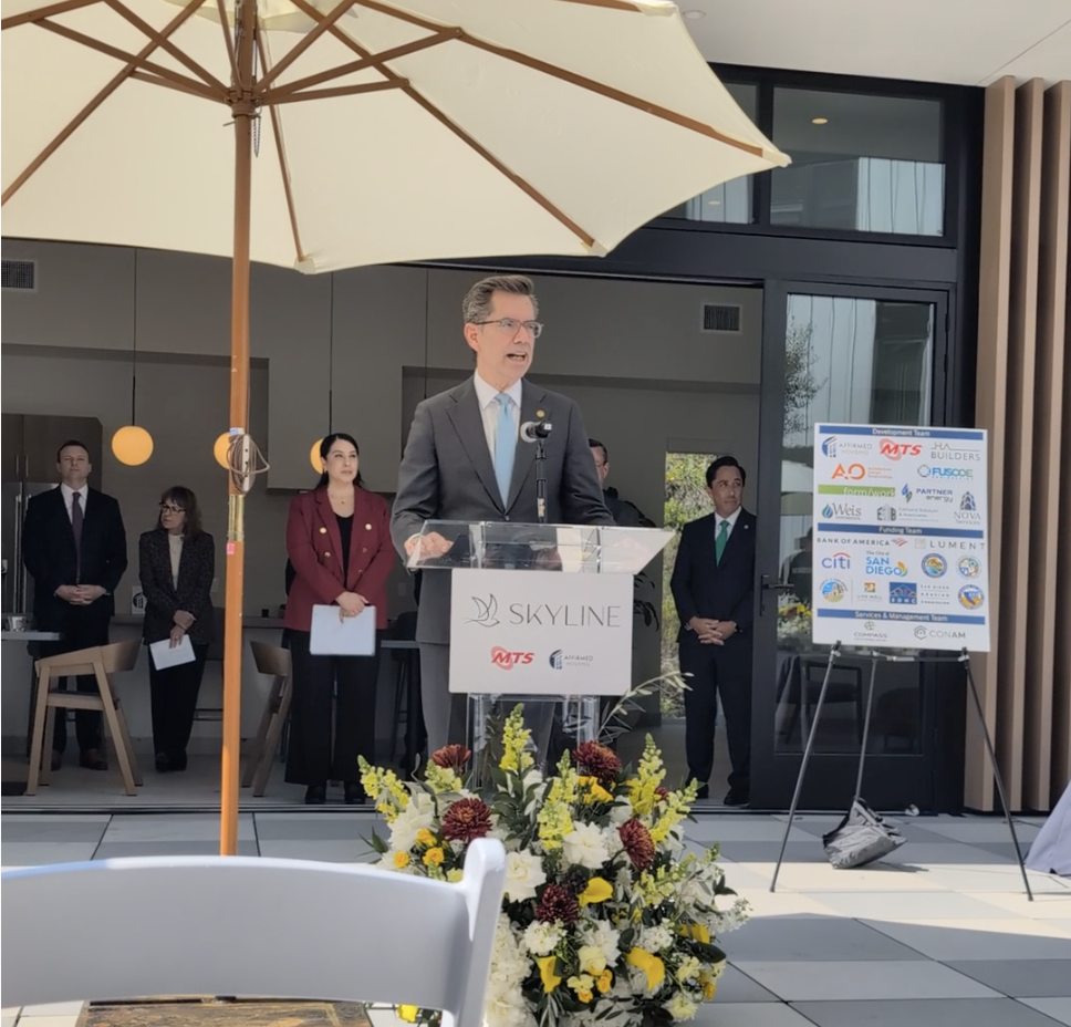 A man in a gray suit and blue tie is giving a speech at a podium with a Skyline sign, outdoors under a large beige umbrella, with a group of professionally dressed people standing behind him. There is a floral arrangement in front of the podium and a signboard displaying logos to the right.