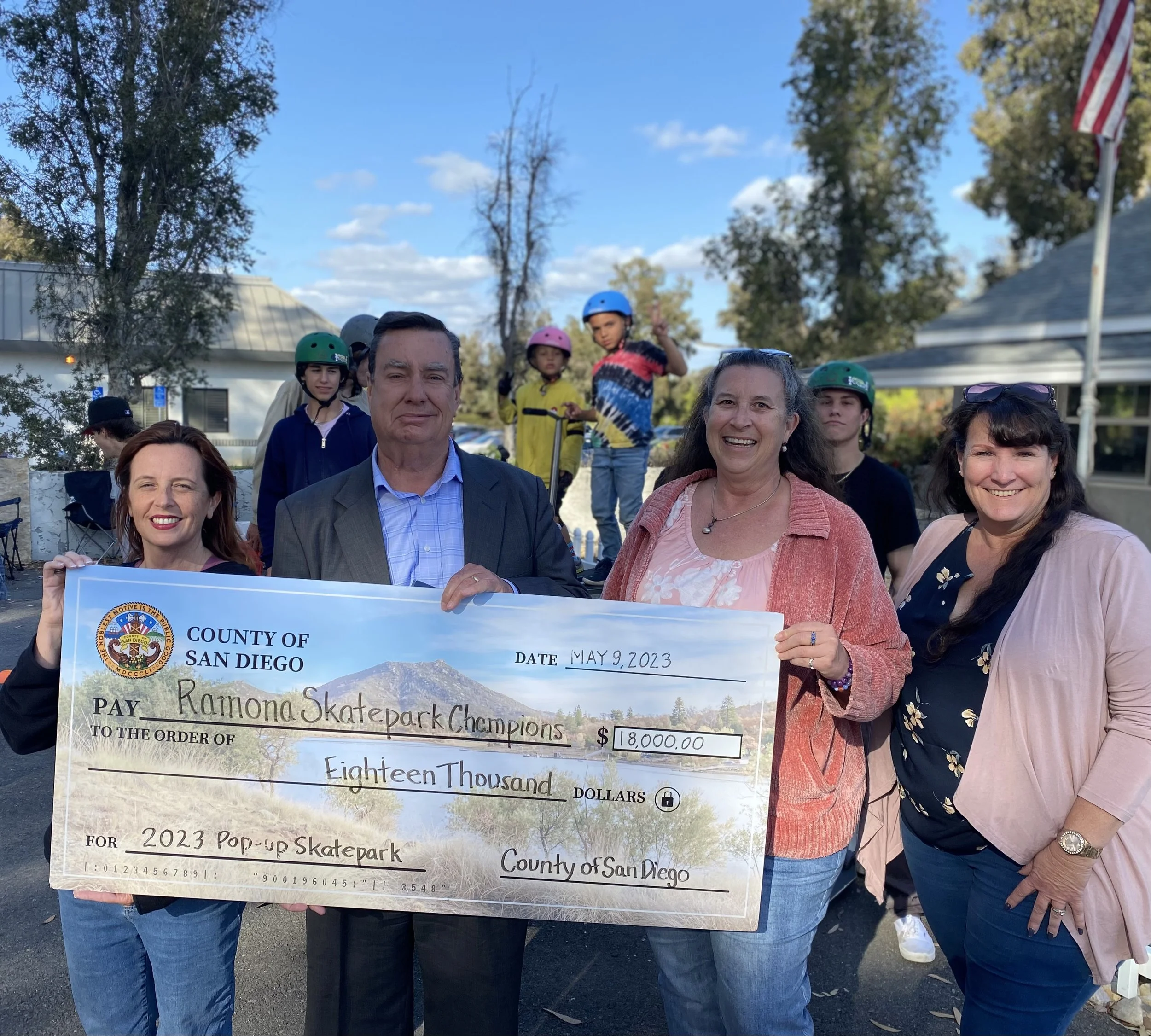 Group of five adults holding oversized check for $18,000 made out to Ramona Skatepark Champions, with children in helmets in the background, outdoors on a sunny day.