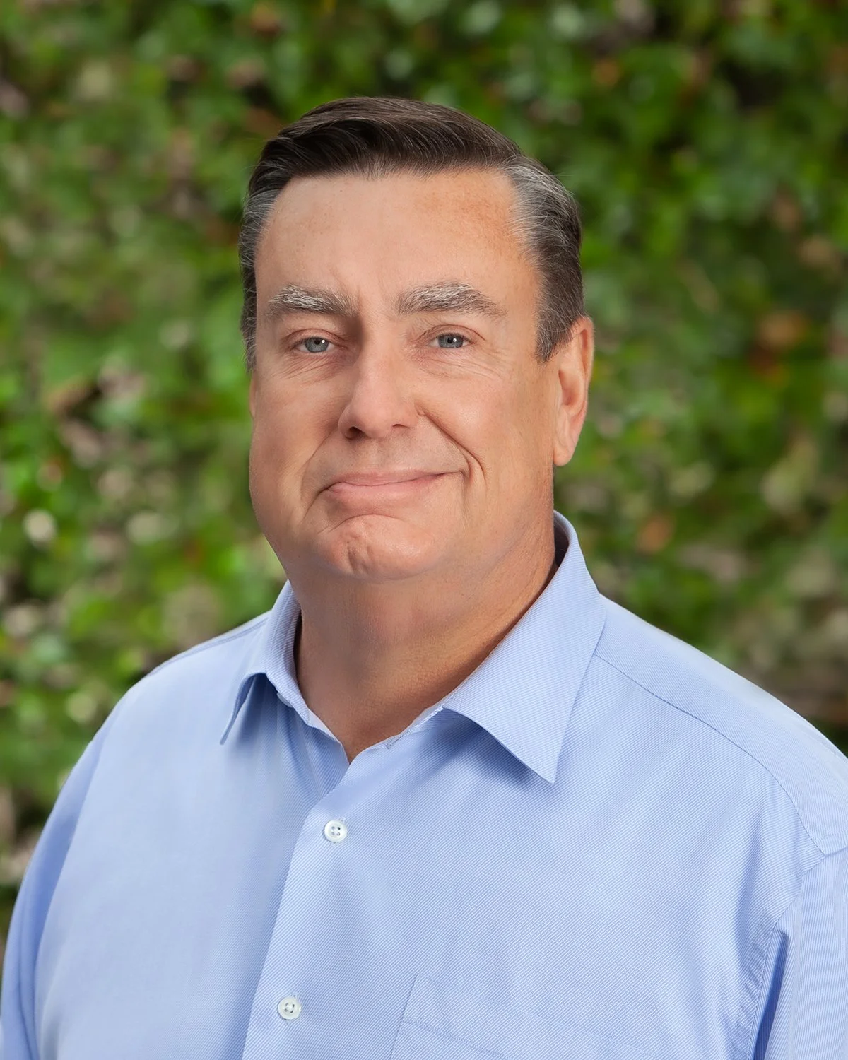 A middle-aged man with dark hair and blue eyes wearing a light blue button-up shirt in front of a green leafy background.