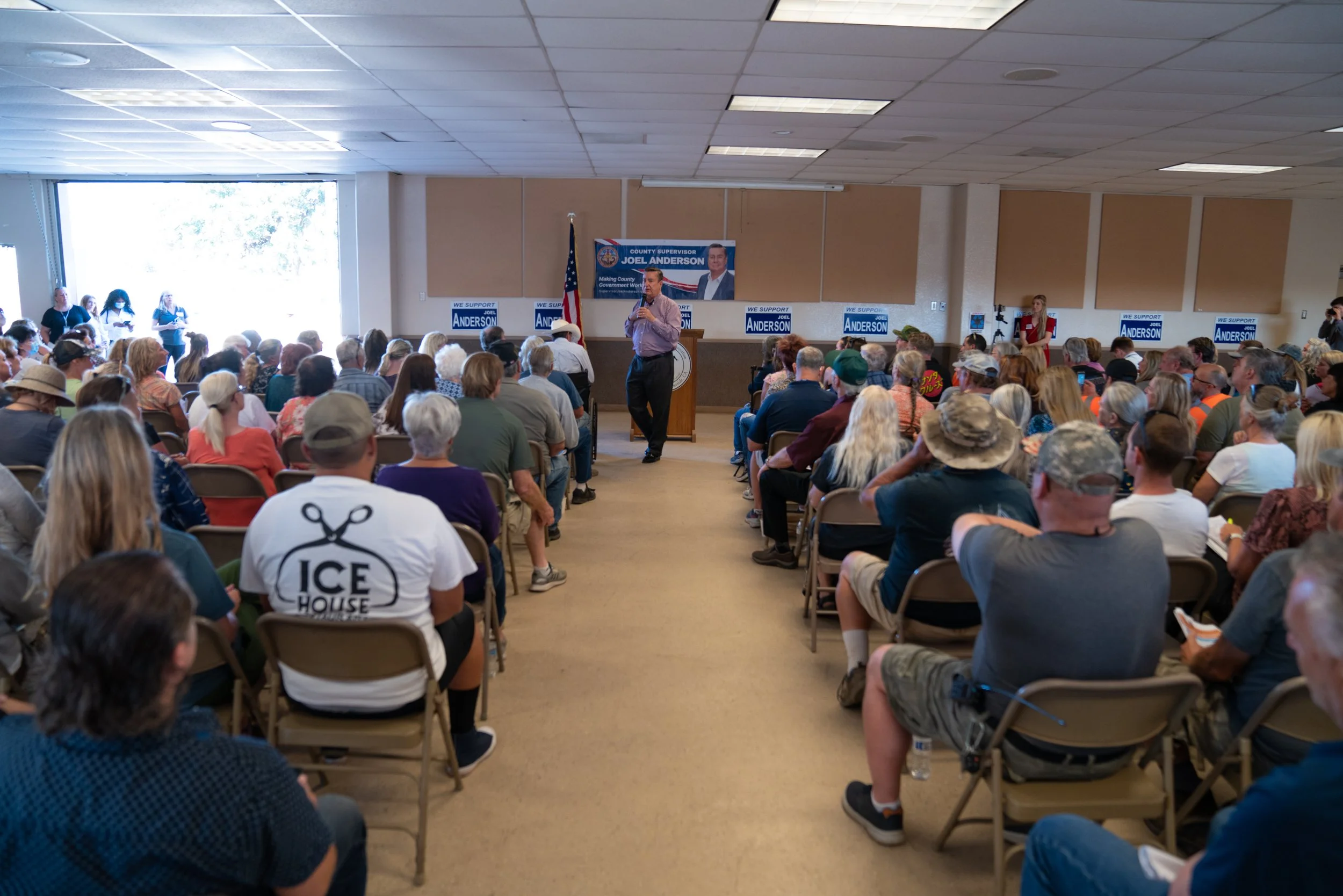 A man is speaking at a podium during a political campaign event in a crowded indoor room with many attendees seated facing him. There are signs and banners for Joel Anderson, with some a supporter wearing a shirt that says 'Ice House' and an American