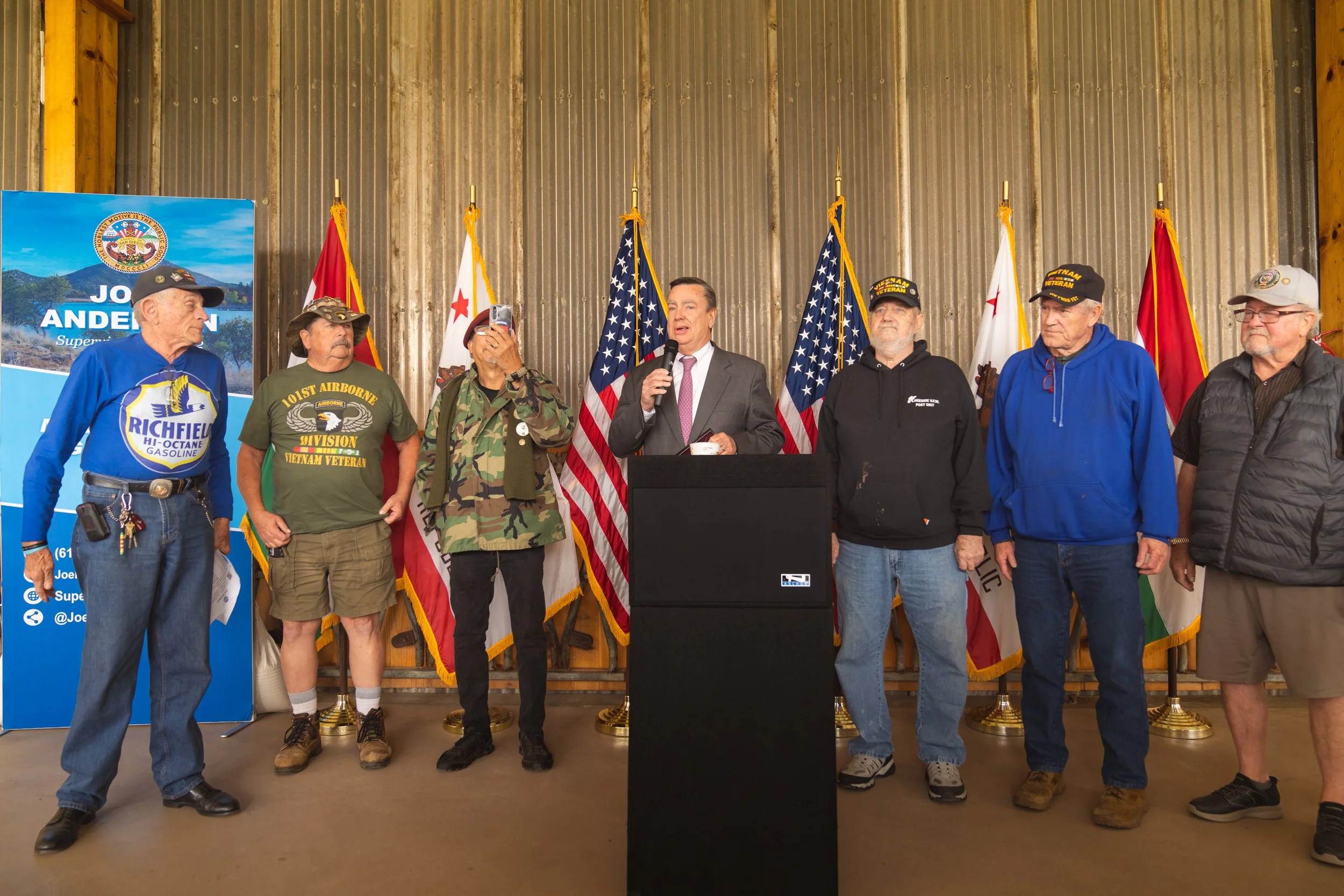 Group of veterans and a man in a suit standing behind a podium with American flags in the background, at a patriotic event.