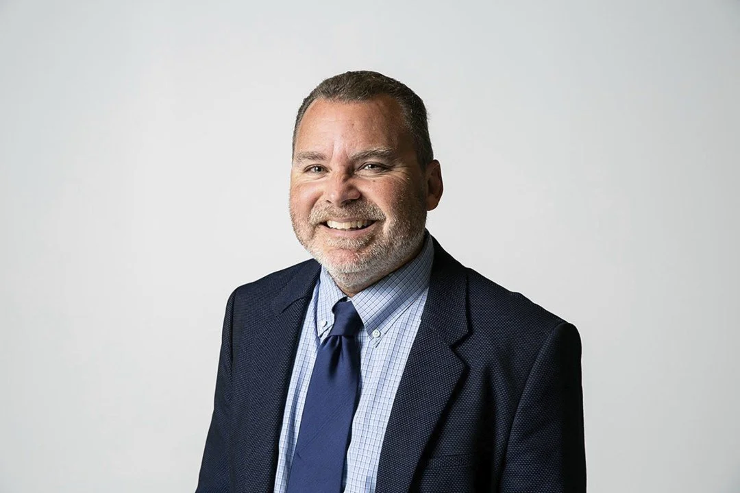 Portrait of a middle-aged man in a dark suit, light blue checkered shirt, and navy tie, smiling in front of a plain light-colored background.