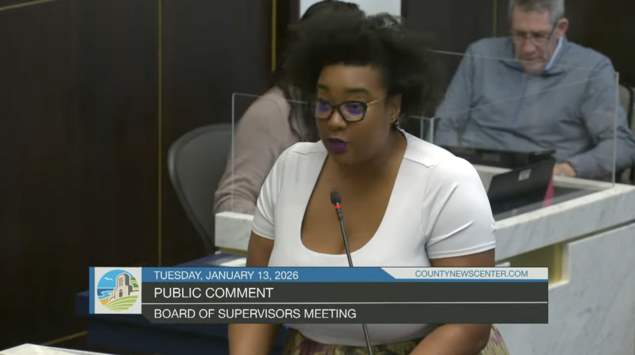 Woman speaking at a public comment during a board of supervisors meeting, with a man in the background using a laptop, date and meeting details displayed on the screen.