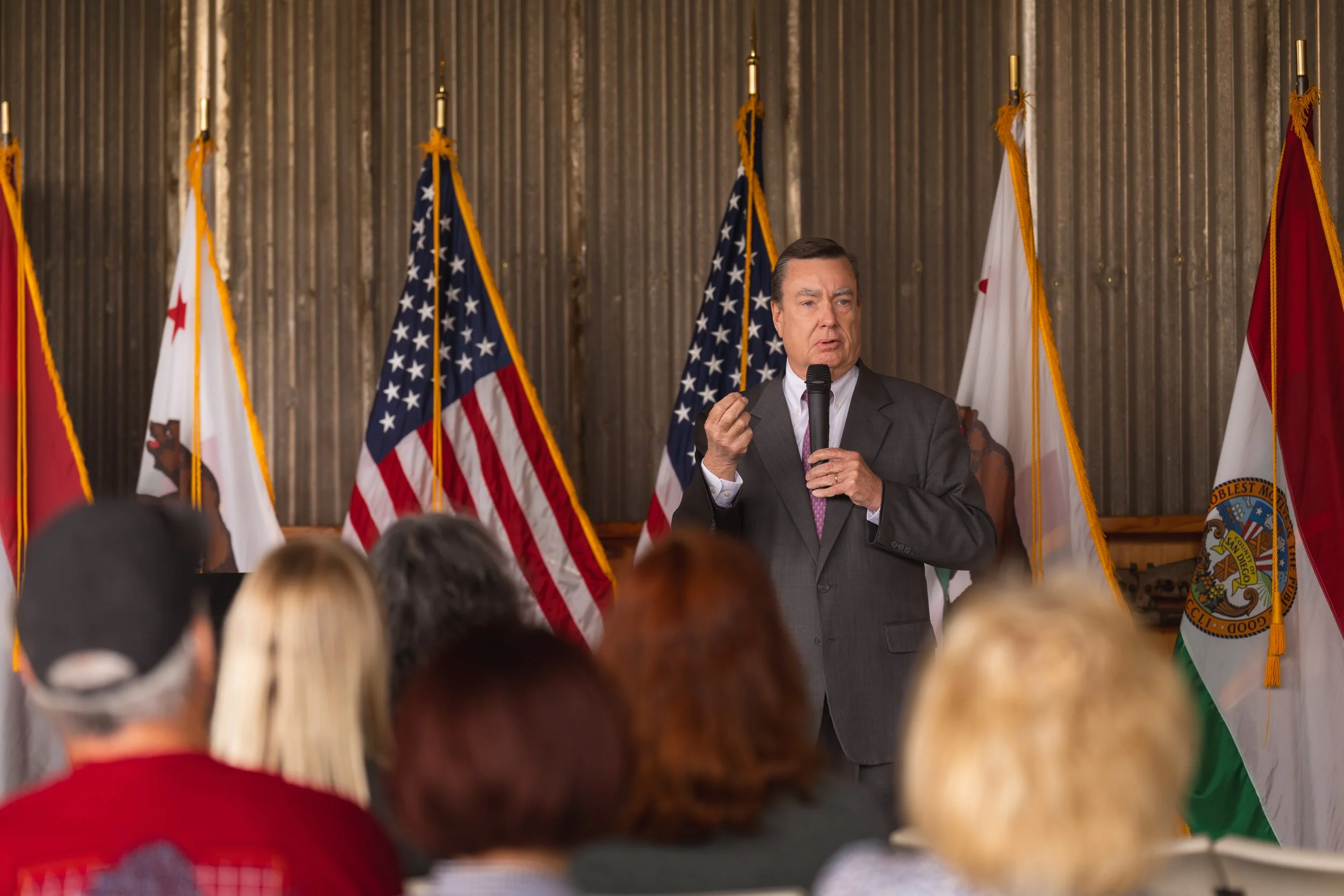 A man in a gray suit speaking into a microphone during a presentation or speech, with American flags and California state flags behind him, and an audience watching.