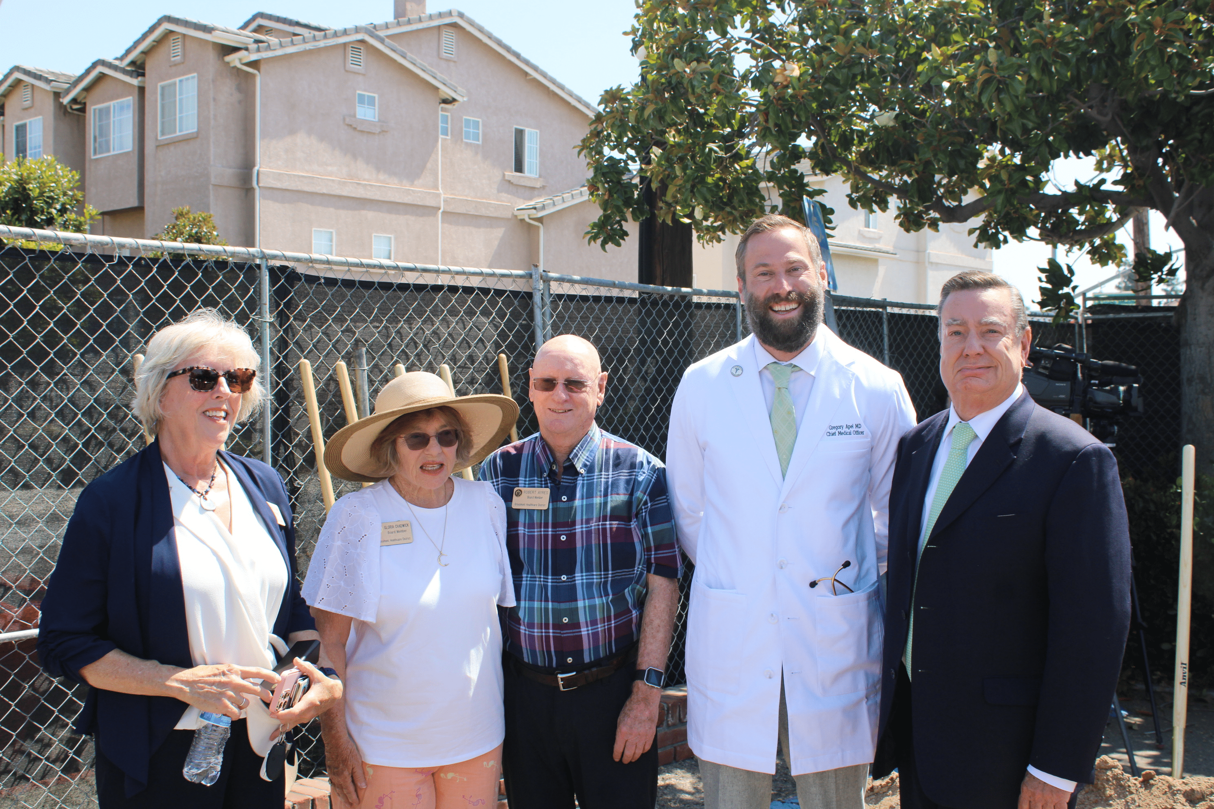Group of five people standing outdoors in front of a chain-link fence and residential buildings, with two women, two men, and a man in a white medical coat, smiling for the photo.
