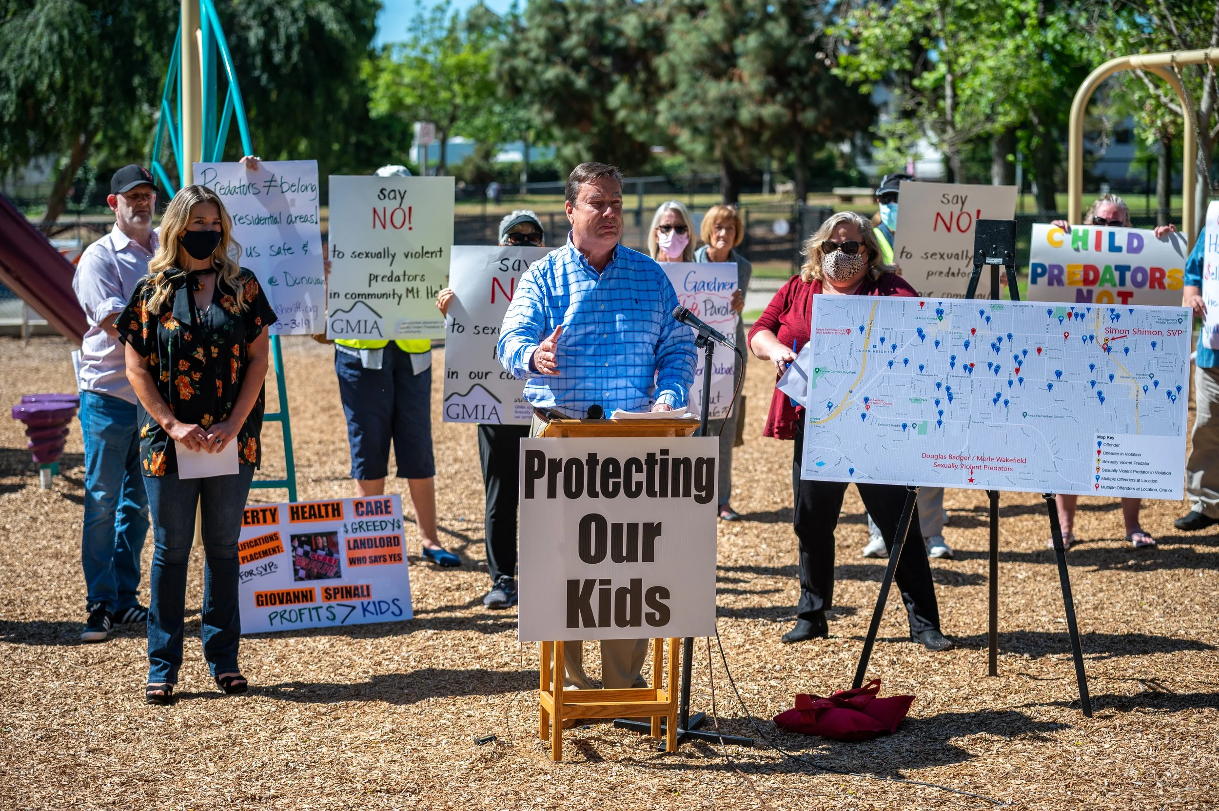 A group of people at a rally, with a man in a blue shirt speaking at a podium that says 'Protecting Our Kids'. Several protesters hold signs with messages like 'say NO to sexually violent predators' and a large map with blue and red pins.