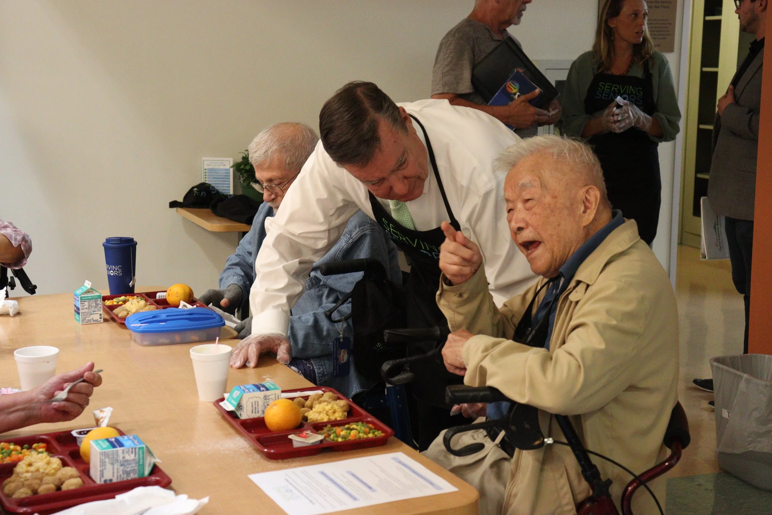 A group of elderly people and caregivers gathered around a table during a meal at a care facility, engaging in conversation.