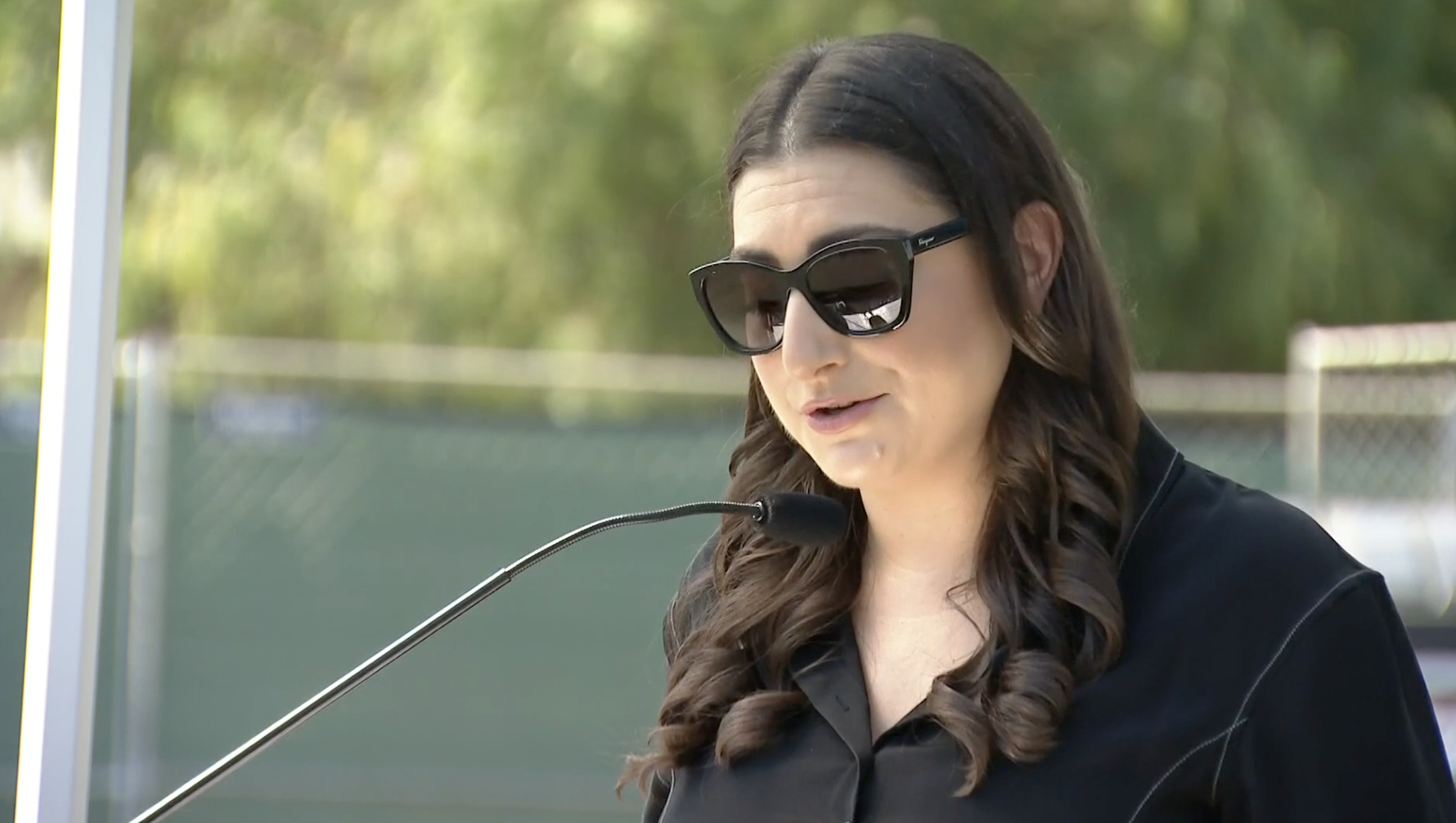 A woman wearing sunglasses and a black shirt speaking into a microphone outdoors.