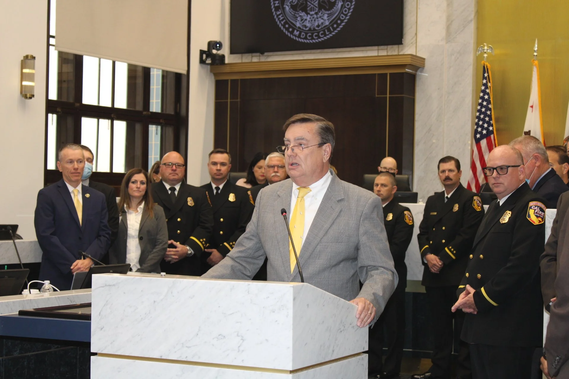 A man in a gray suit and yellow tie is speaking at a podium in a formal meeting or government setting, with several uniformed police or fire officers and other officials standing behind him, along with flags and an official emblem on the wall.
