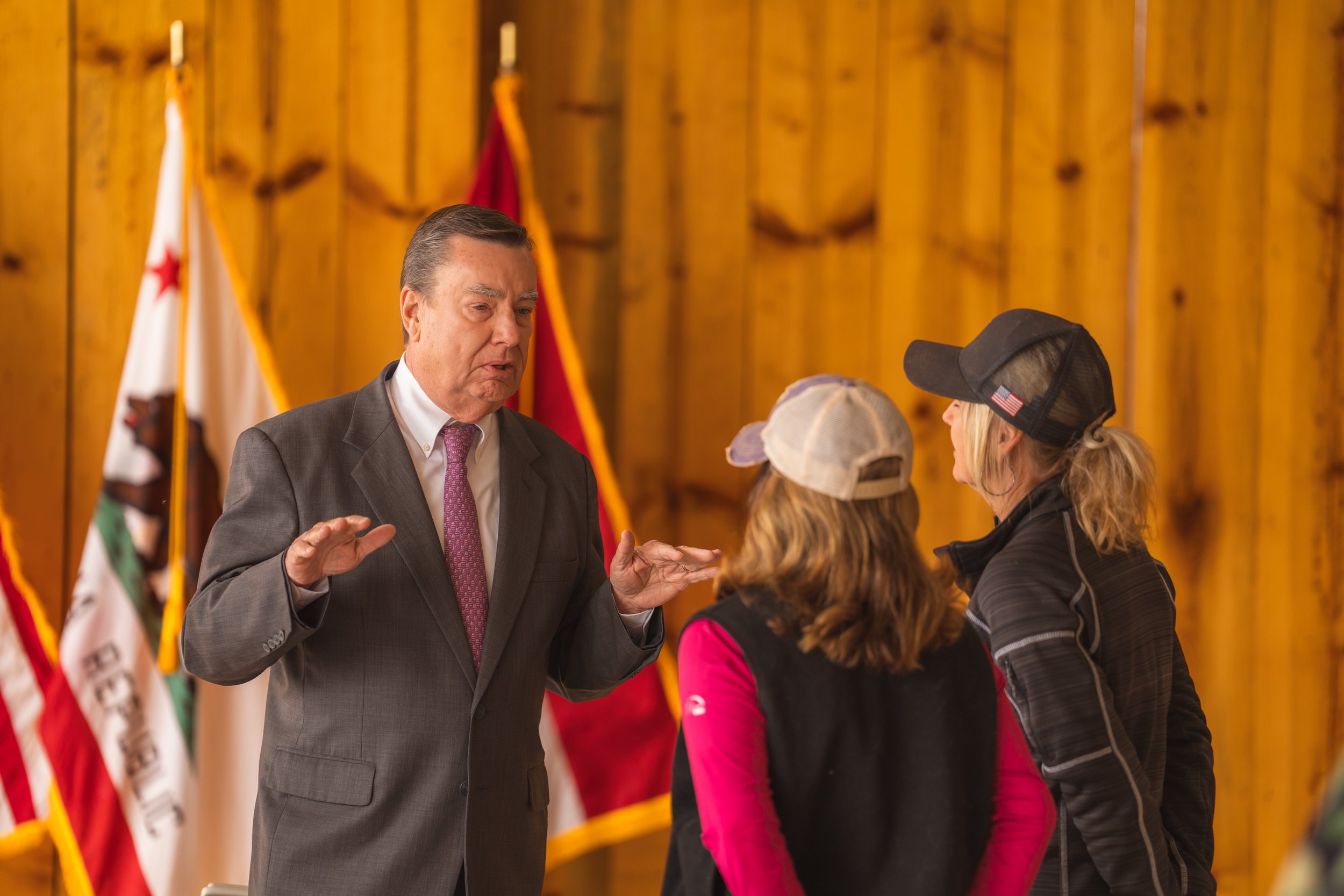 A man in a suit talking to two women wearing casual clothes and baseball caps, inside a wooden room with multiple American flags in the background.
