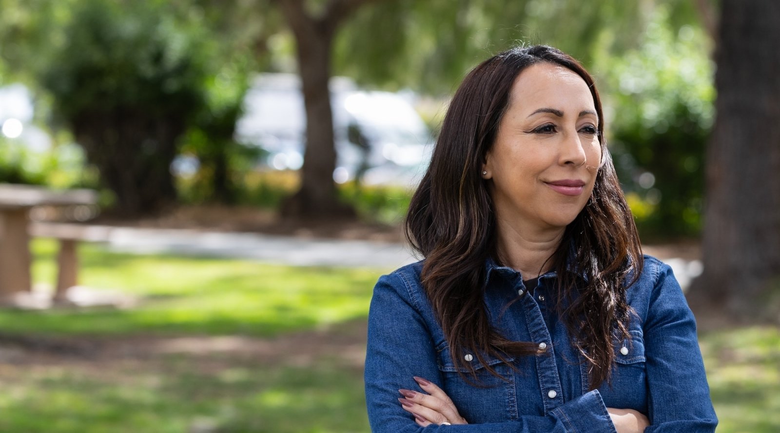 A woman with dark brown hair in a forested park with trees and green grass, smiling with arms crossed, wearing a blue denim jacket.