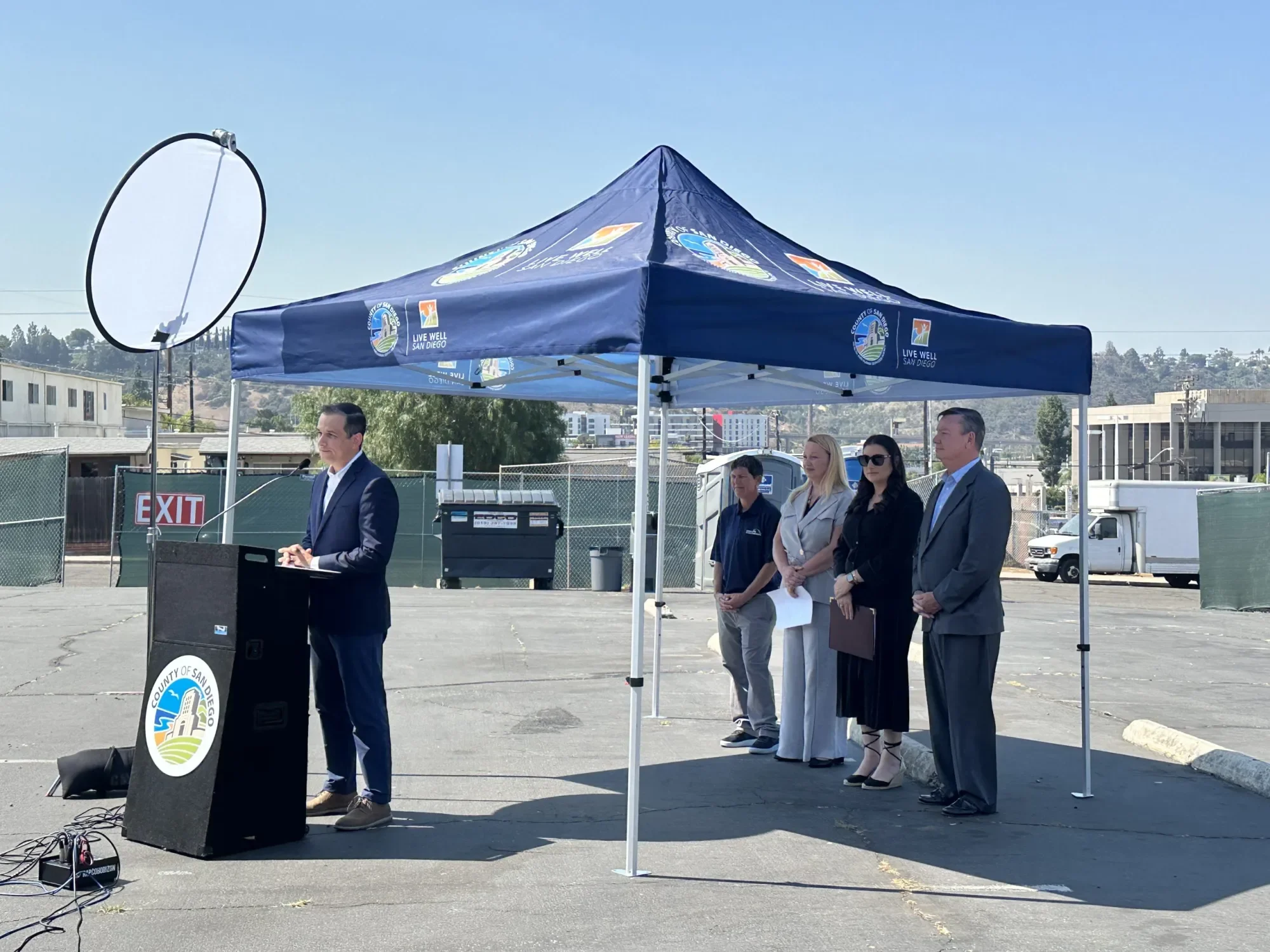 A man in a suit gives a speech at a podium with the County of San Diego logo. Four people stand behind him under a blue canopy with County of San Diego logos, outdoors in a parking lot with a fence, buildings, and a clear sky.