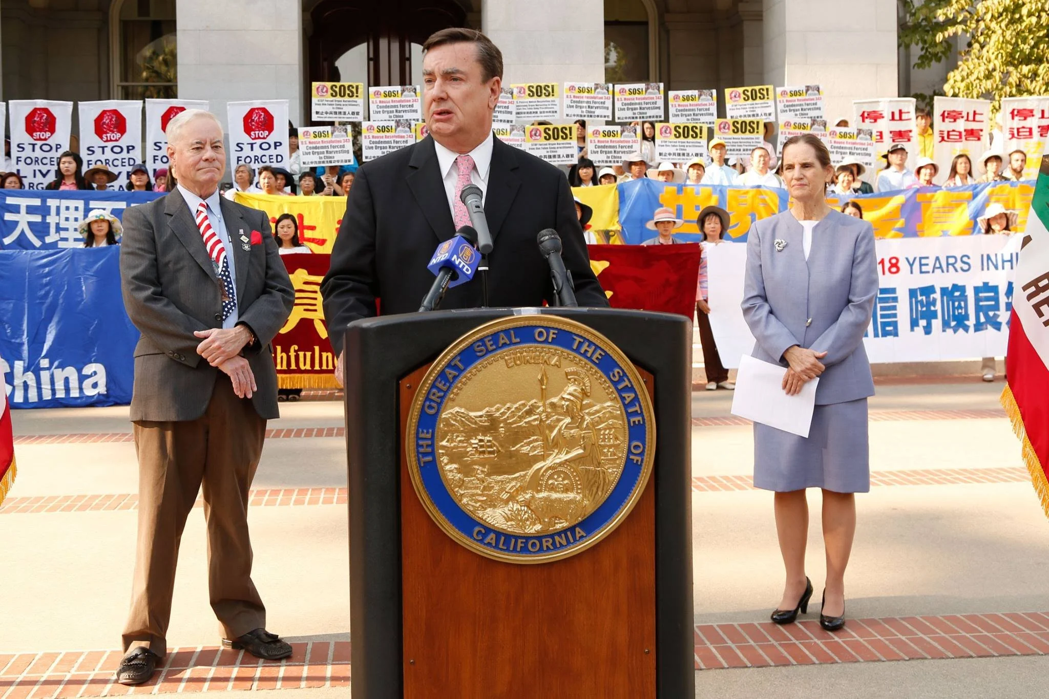 A man in a dark suit and pink tie speaks at a podium with the California state seal, flanked by two people in suits, with a crowd holding protest signs in the background.