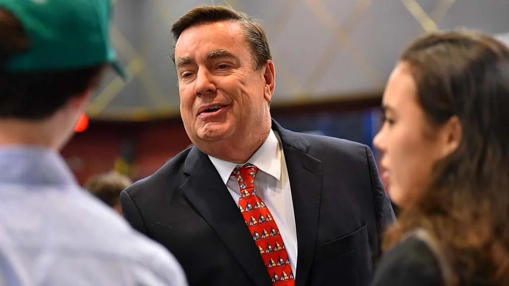 A middle-aged man in a black suit, white shirt, and a red tie with sailboats, talking to two younger people in a social setting.