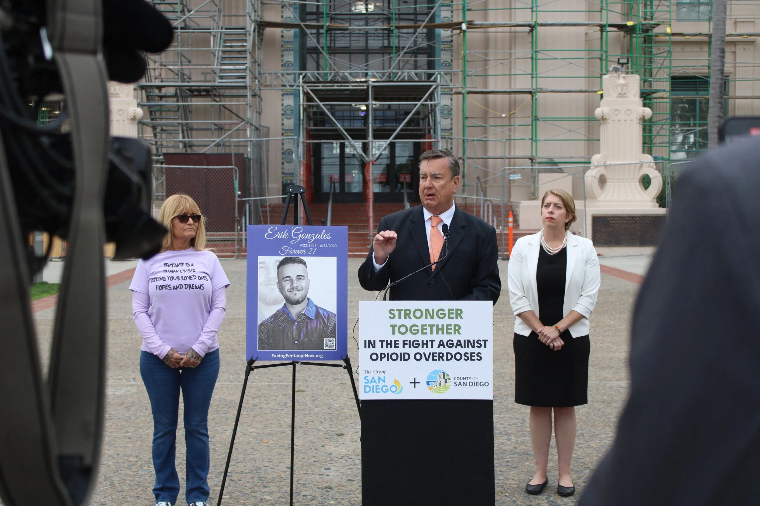A man in a suit speaking at a press conference with a sign that reads 'Stronger Together in the Fight Against Opioid Overdoses,' two women standing behind him, one wearing sunglasses and a purple shirt and the other in a white blazer, with a poster o