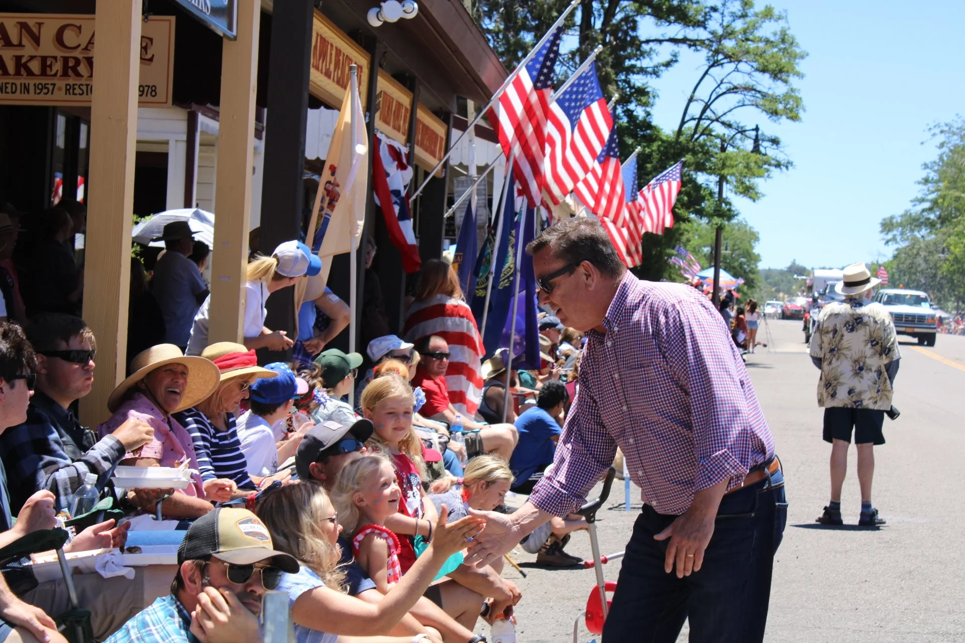 A man shaking hands with a young girl sitting on a curb during a parade, with a crowd of spectators, American flags, and flags of various states and organizations visible in the background on a sunny day.