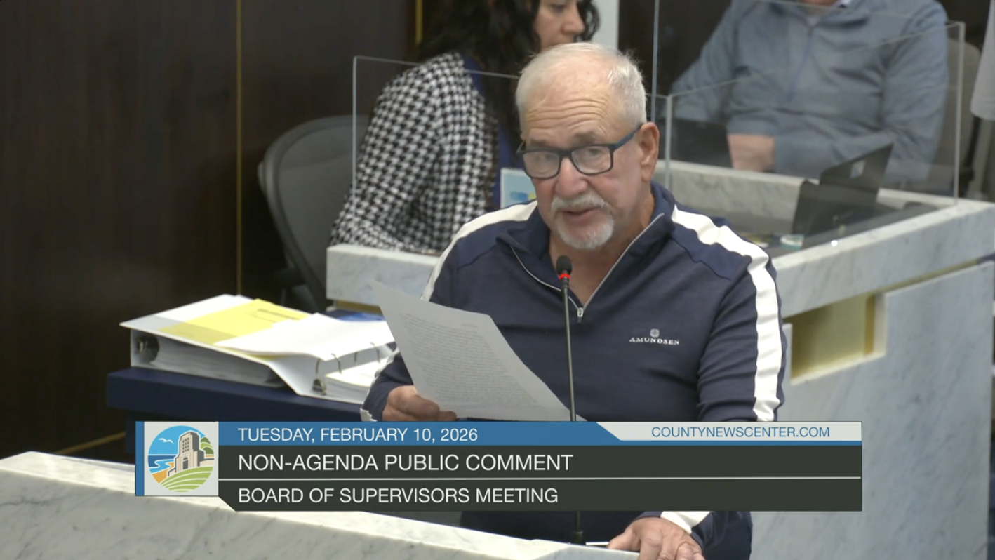 A man with glasses and white hair speaking at a microphone during a public comment at a board of supervisors meeting, reading from a paper. There are other people in the background and paperwork on the table.