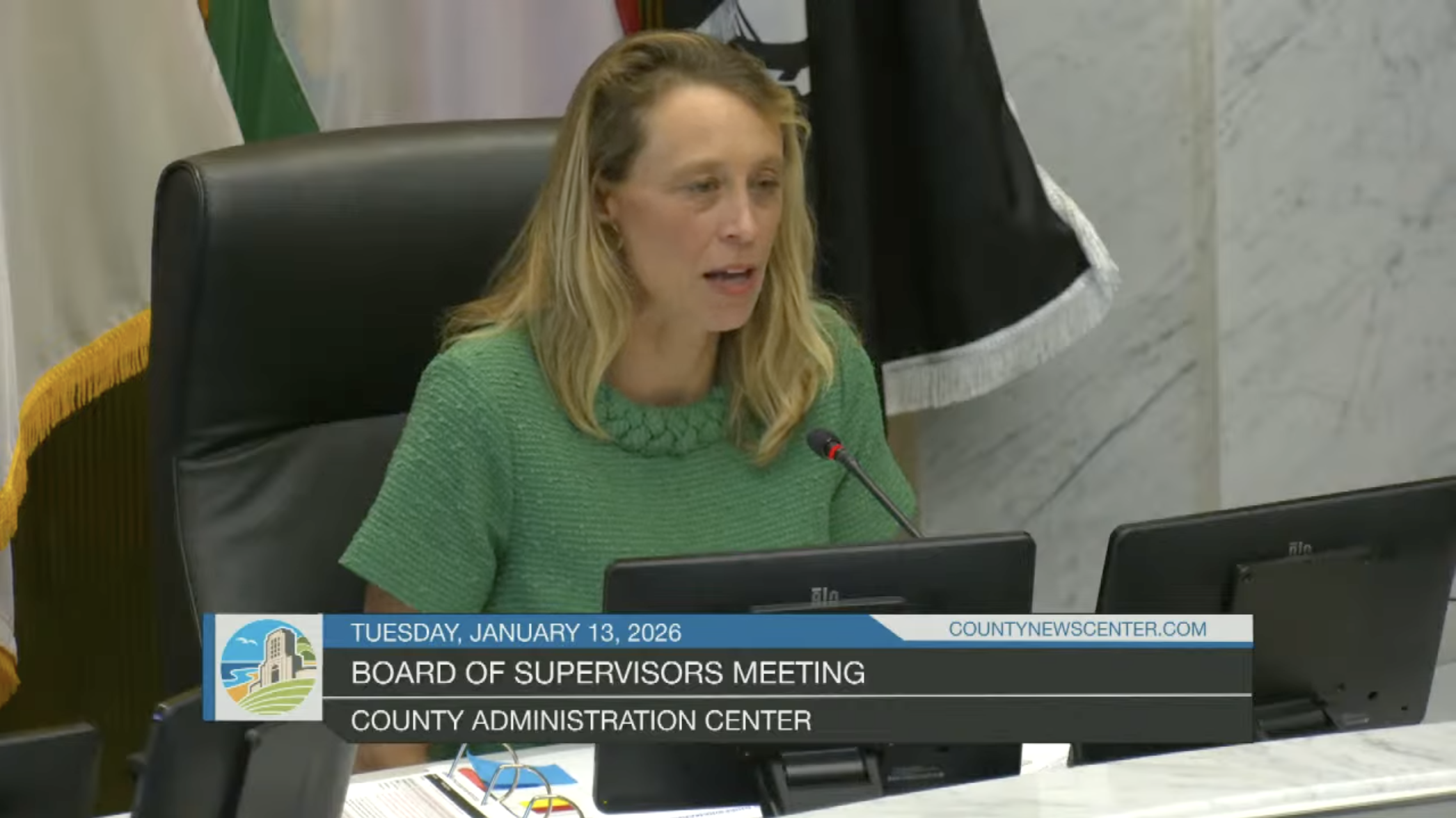 A woman with blonde hair wearing a green shirt speaking into a microphone at a county supervisors meeting in the County Administration Center. There are computer monitors in front of her and flags in the background.