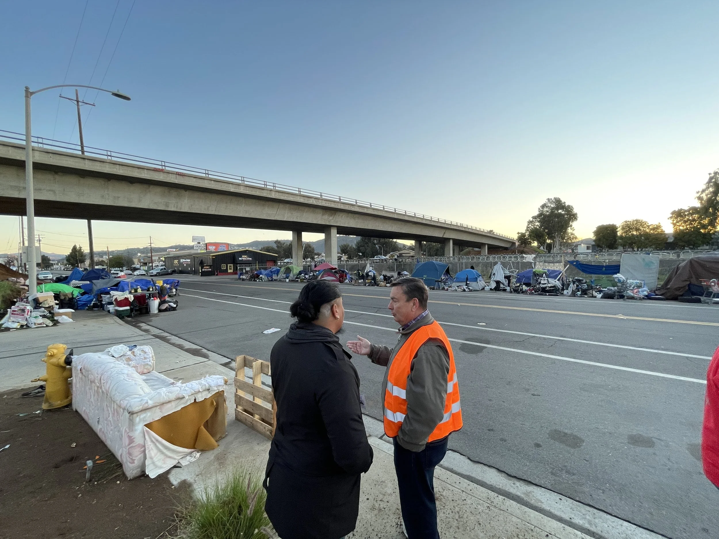 Two people having a conversation on a city street with numerous tents and belongings in the background, suggesting a homeless encampment along the sidewalk.