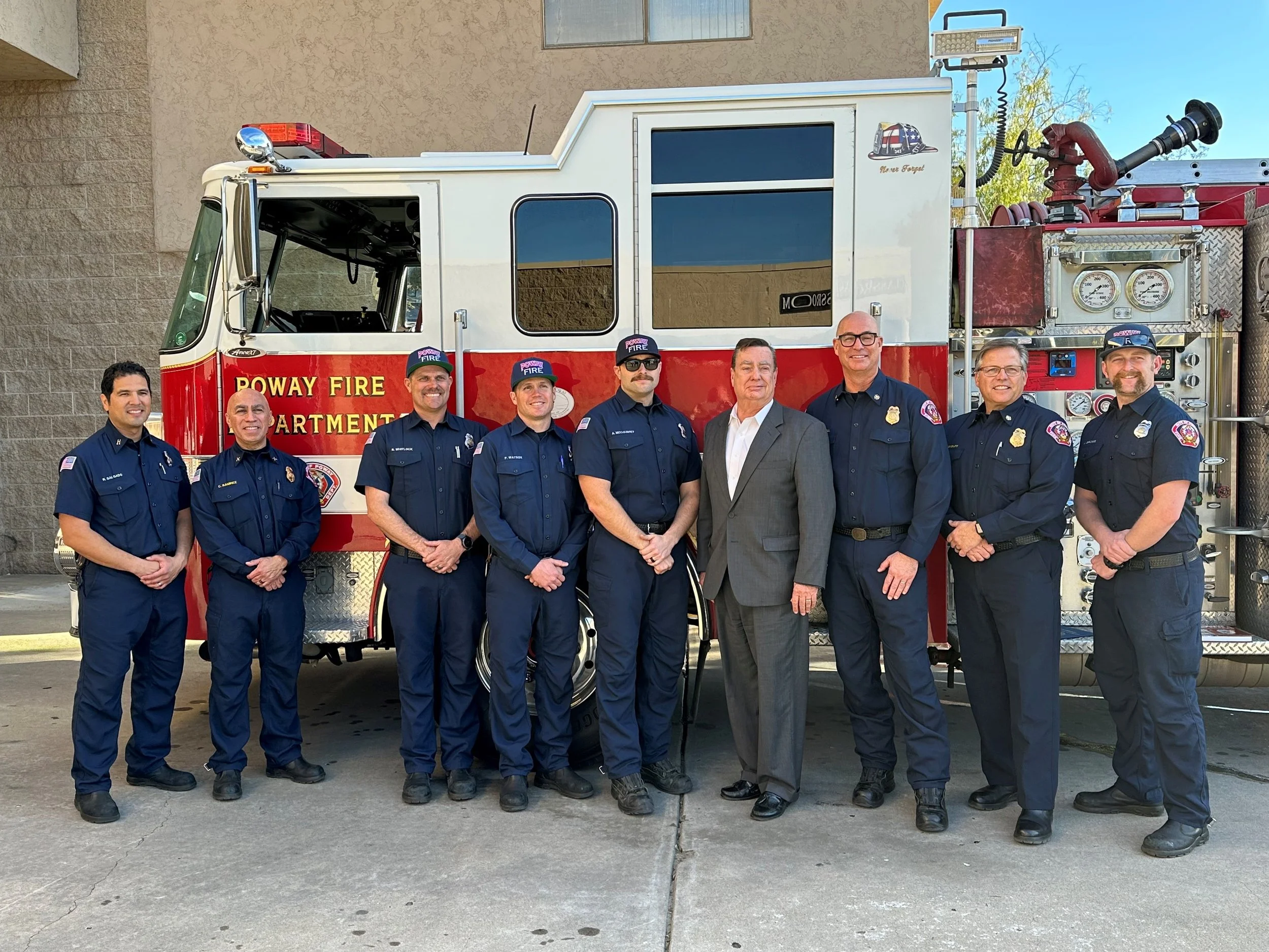 Group of nine firefighters and a man in a suit standing in front of a red and white fire truck marked 'Poway Fire Department'.