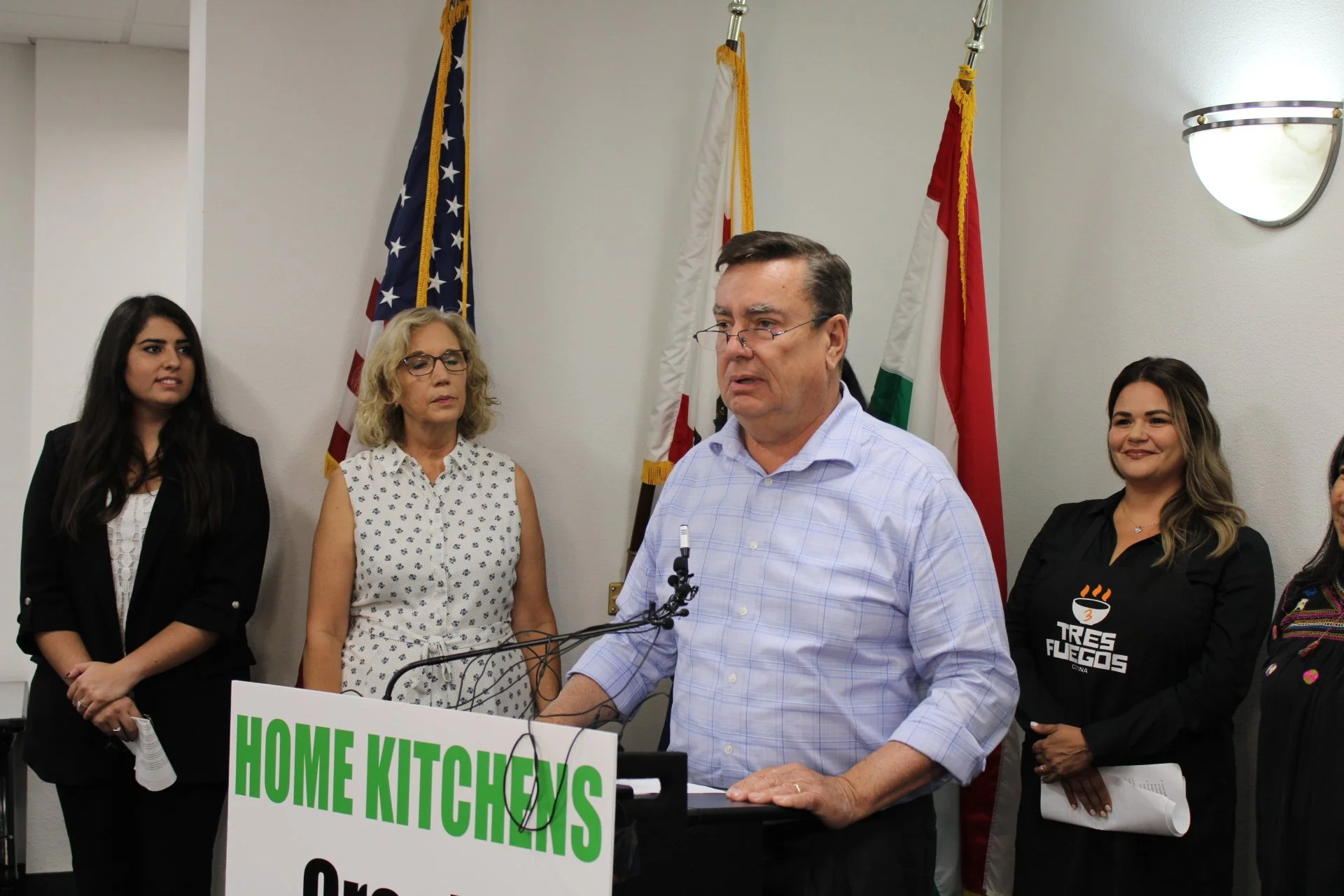 A man giving a speech at a podium labeled 'Home Kitchens' with four women standing behind him, in front of American and other flags, in a room with white walls and a wall-mounted light.