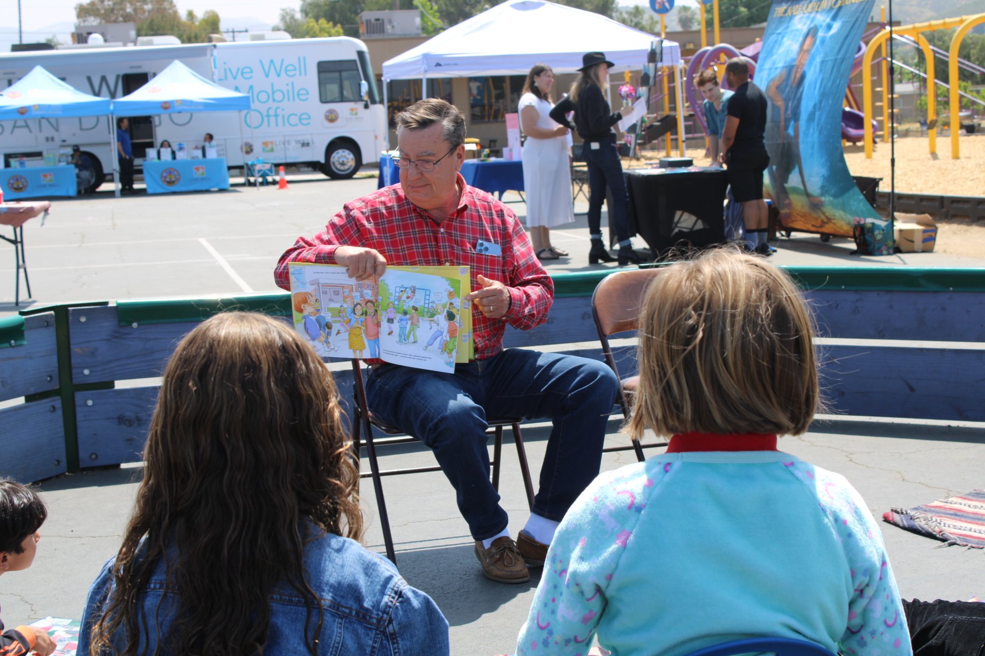 A man reading a children's book to a group of kids seated outside at an event with booths and tents in the background.
