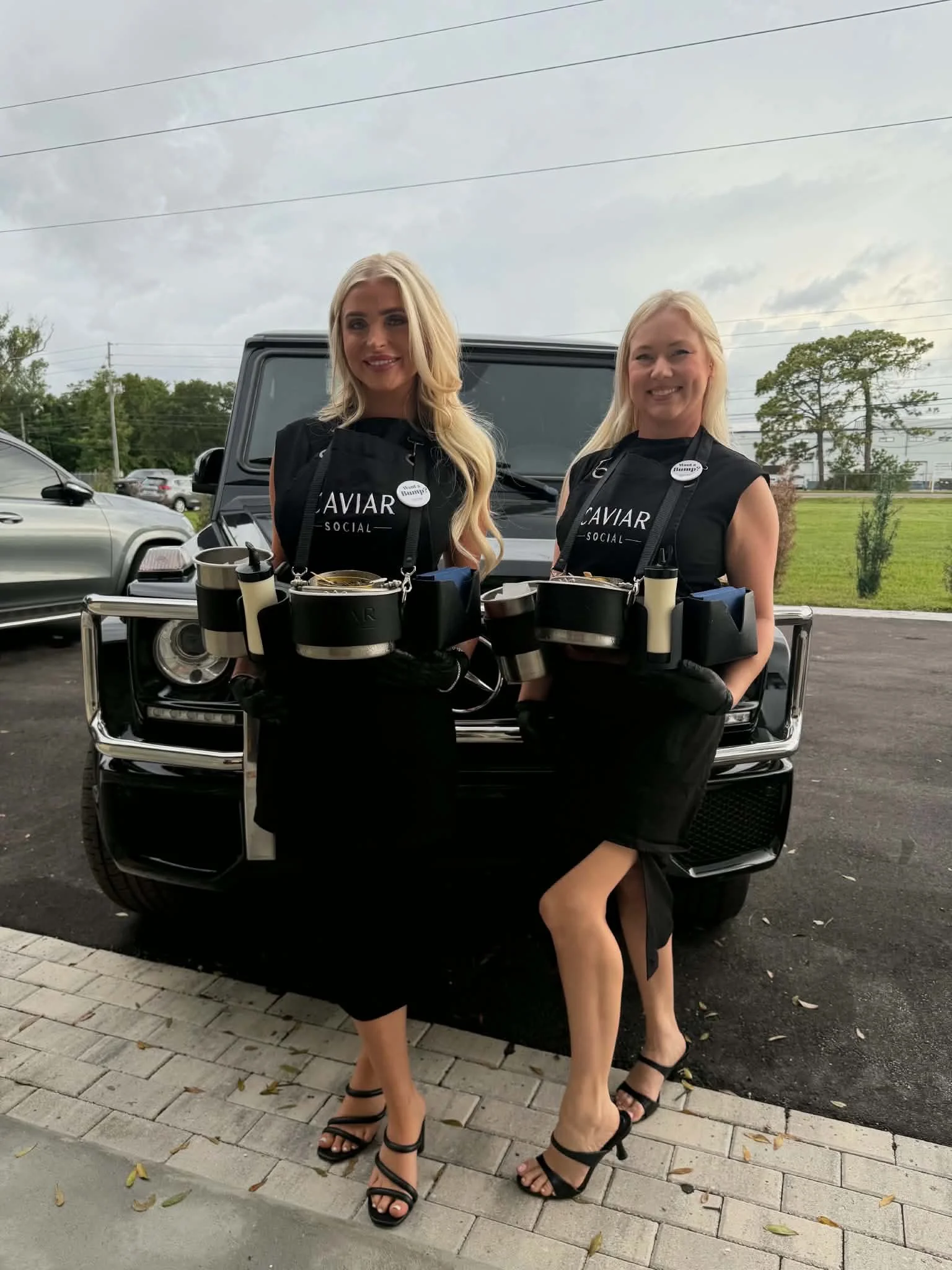 Two women dressed in black, wearing high heel shoes, standing next to a black vehicle outdoors on a cloudy day. One woman has long blonde hair and the other has shoulder-length blonde hair, both smiling.
