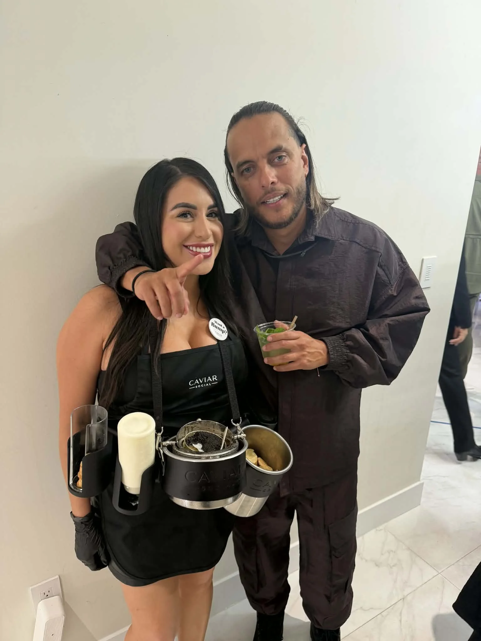A woman smiling and pointing at a man with long hair, both standing against a plain white wall. The woman is wearing a black apron from Caviar Social with condiments and tools attached. The man is holding a drink and wearing a dark jacket.