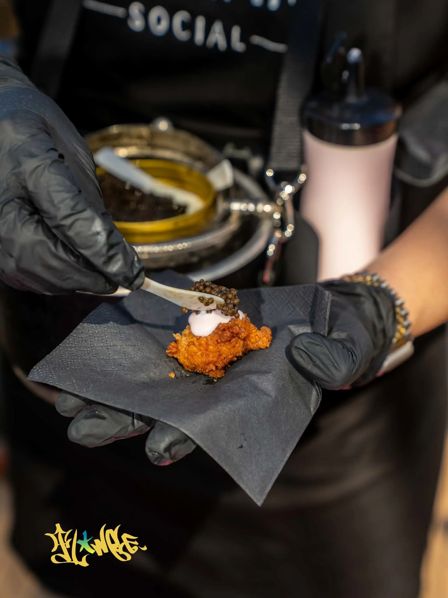 Person wearing black gloves holding a piece of fried chicken topped with small round black caviar on black paper