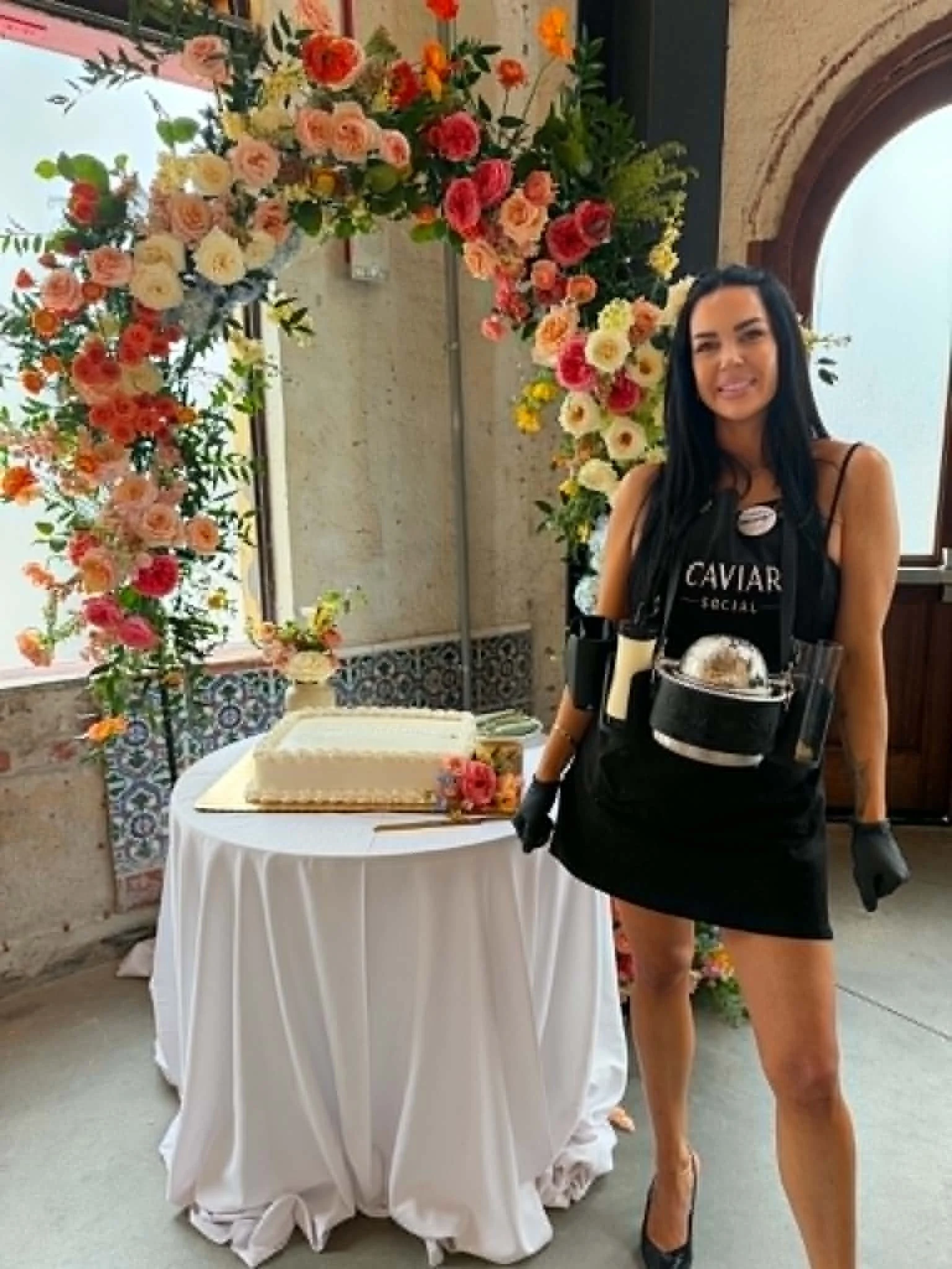 A woman dressed in black serving staff attire stands next to a dessert table at a celebration, with colorful flowers and a cake in the background.