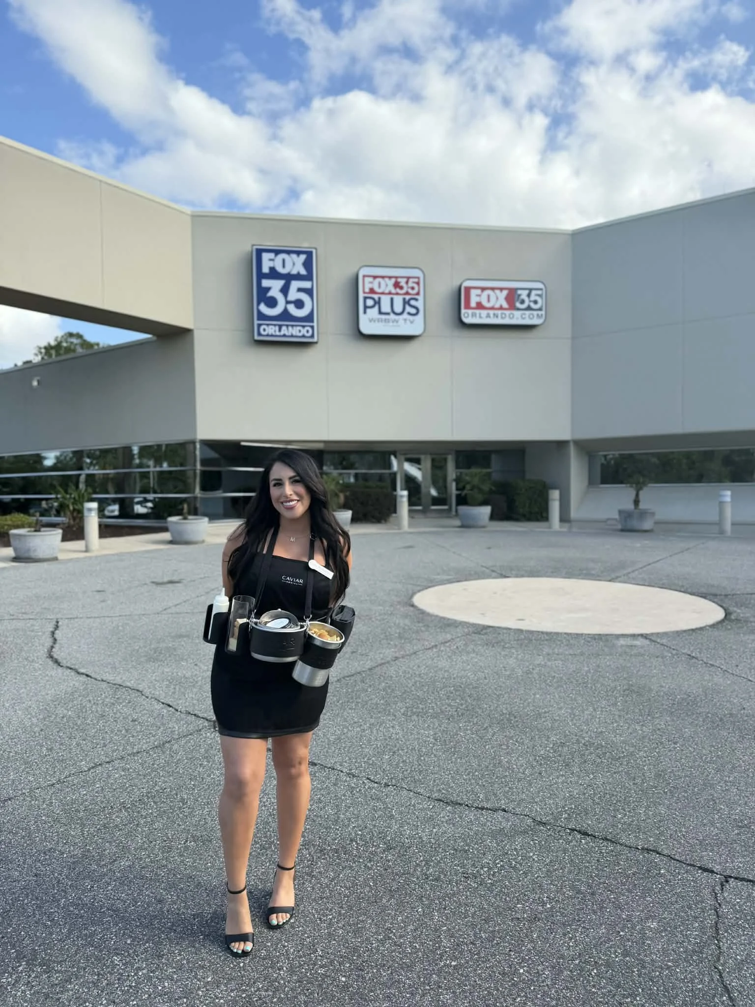 A waitstaff woman holding a tray with food and drinks, standing outside the Fox 35 Orlando building on a sunny day with a partly cloudy sky.