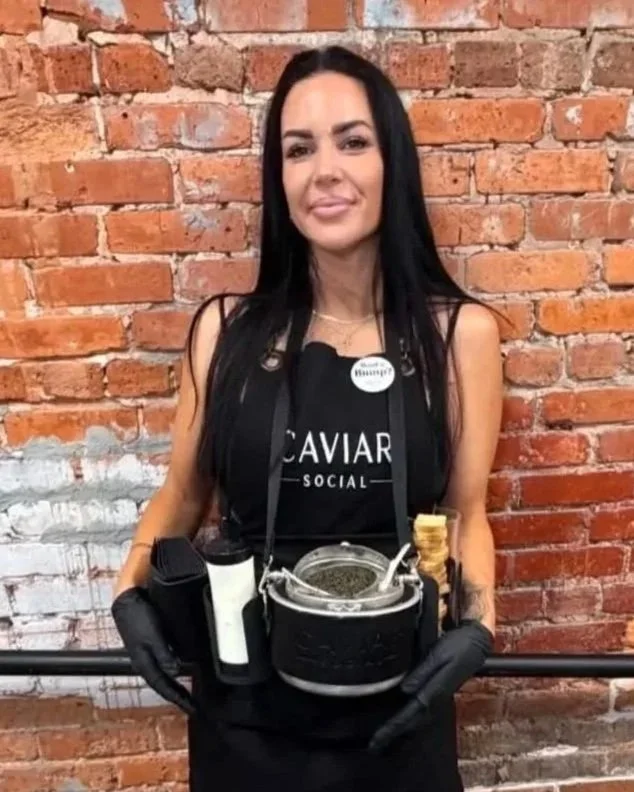 A woman with long black hair and a black apron standing against a brick wall, holding a tray of caviar and chips, wearing black gloves and a name badge that says 'Tiffany'.