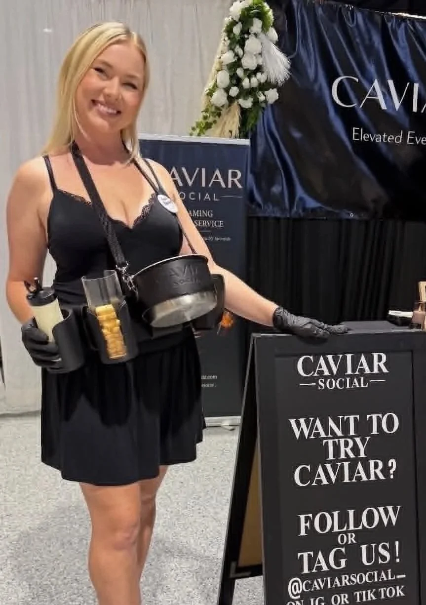 A woman smiling at a promotional booth for caviar social, dressed in black, holding containers of caviar. The booth has a large sign asking 'Want to Try Caviar? Follow or Tag Us!' with candles, white flowers, and decorative feathers nearby.