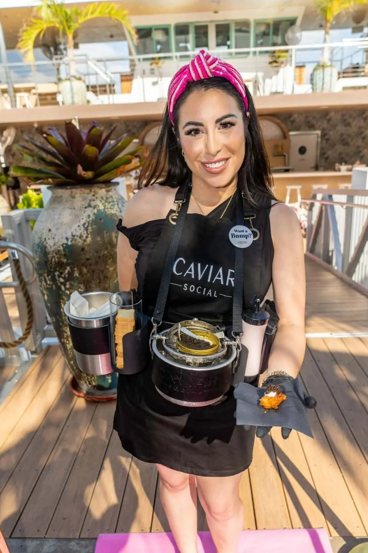 A smiling woman with dark hair wearing a pink striped headband, black apron with 'Caviar Social' logo, holding a tray with caviar, standing on a wooden deck outdoors with a large potted plant and a building in the background.