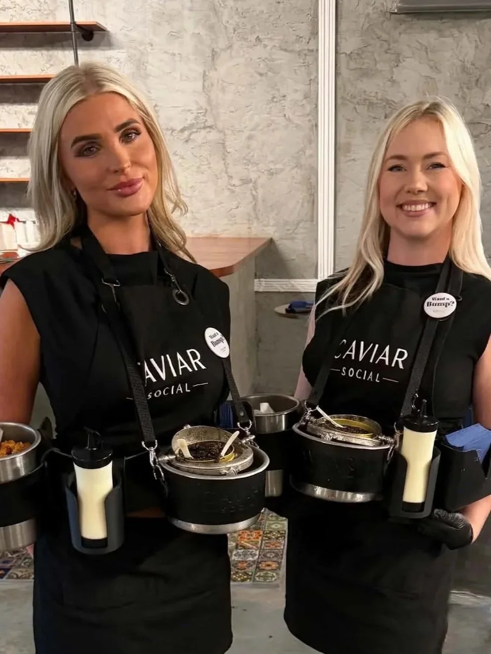 Two women standing side by side wearing black aprons with 'Caviar Social' printed on them, smiling, holding sauce containers and serving trays in a modern kitchen or cafe setting.