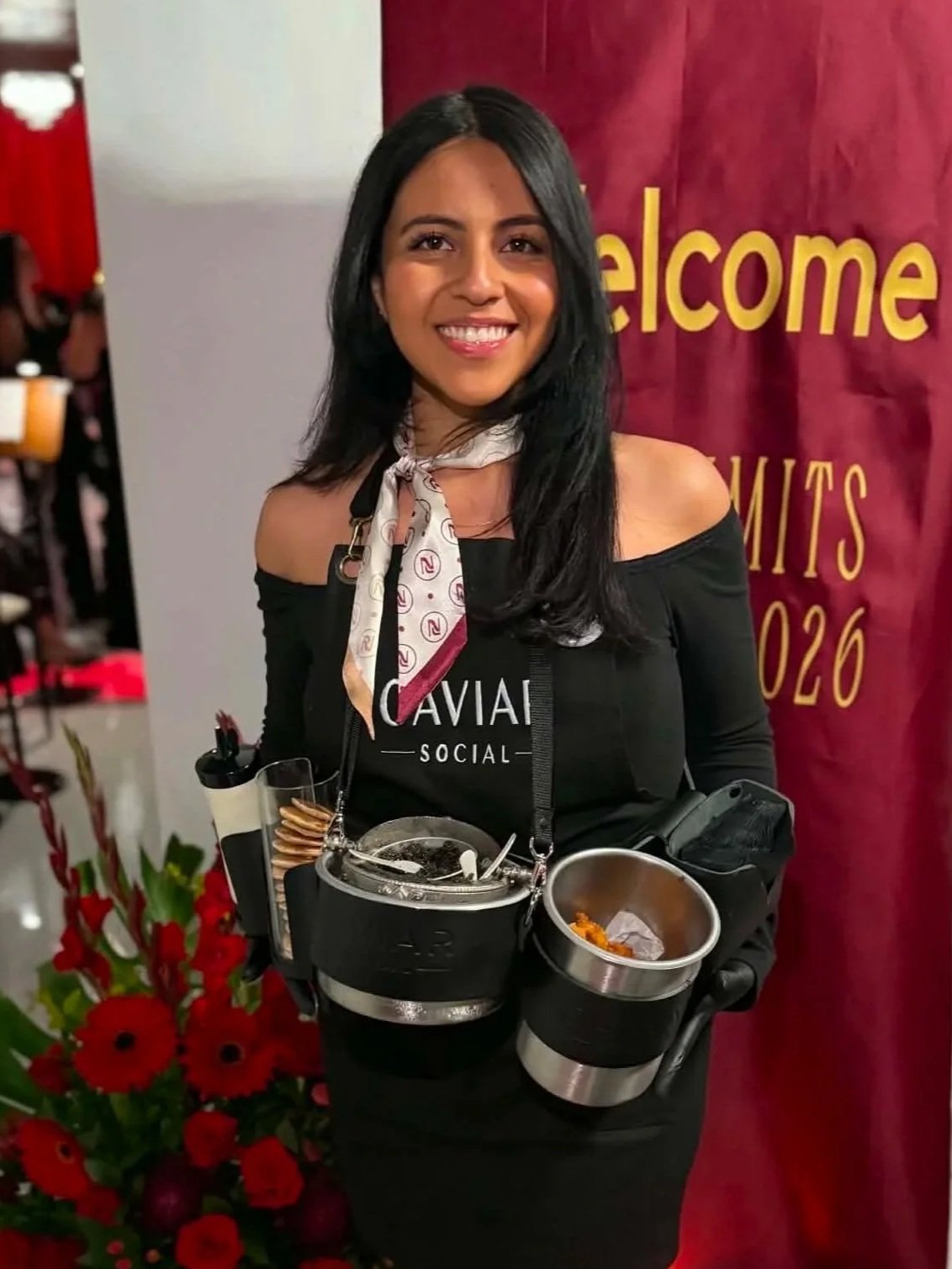 A smiling woman with black hair, wearing off-the-shoulder black dress and a white scarf with red patterns, standing in front of a red and yellow "welcome" sign. She is holding a tray of food and surrounded by red flowers.
