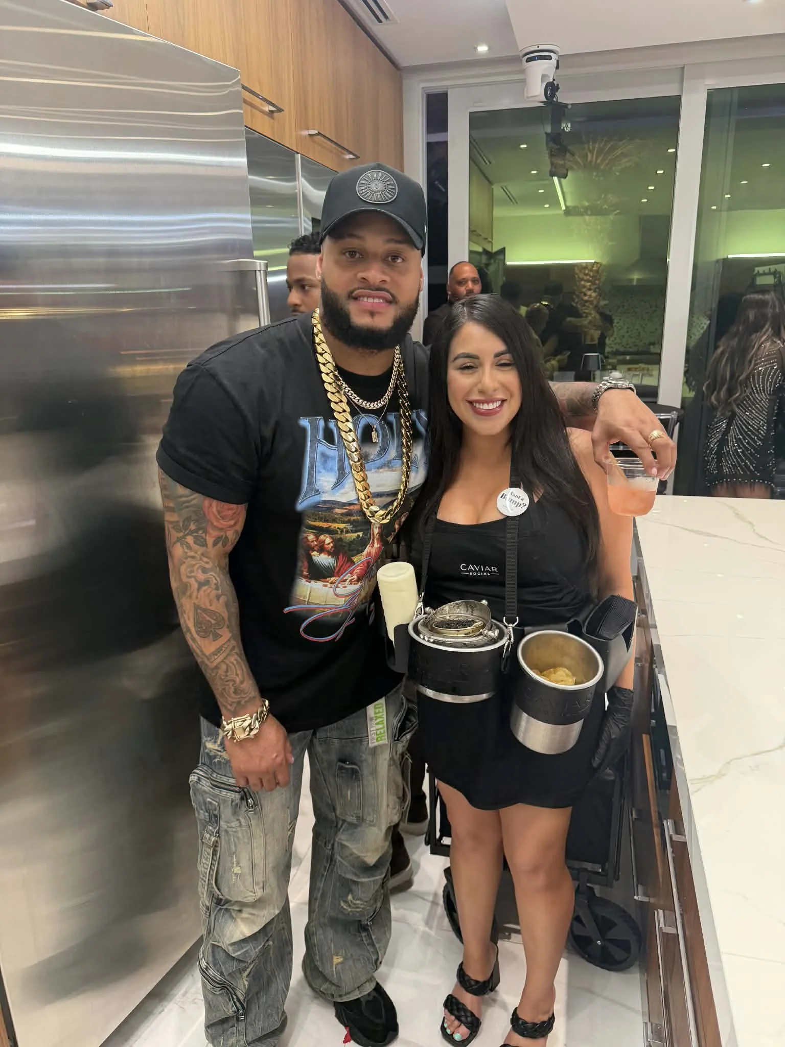 A man and a woman posing Caviar social event, with the man wearing a black t-shirt, cap, and lots of gold jewelry, and the woman dressed in black with a caviar service around her neck, smiling at the camera, standing in a modern indoor space.